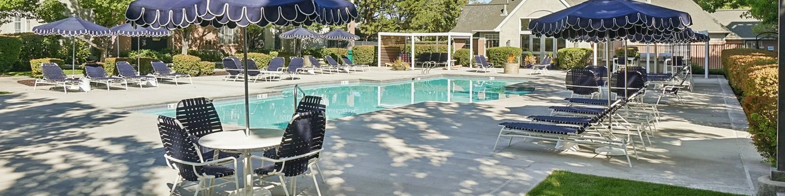 Elegant outdoor pool area with stylish blue umbrellas and sun loungers, surrounded by lush greenery and a luxurious residential building.