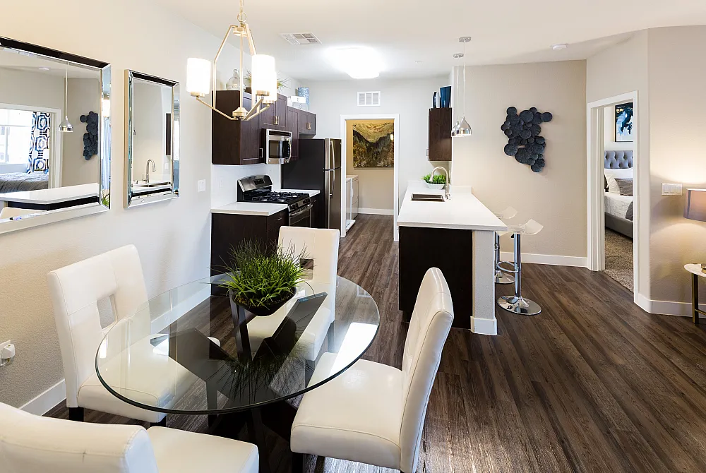 Modern apartment dining and kitchen area with sleek white furniture, stylish lighting, and a warm, inviting atmosphere. High-end kitchen appliances and a glimpse into a cozy bedroom create a welcoming home environment.