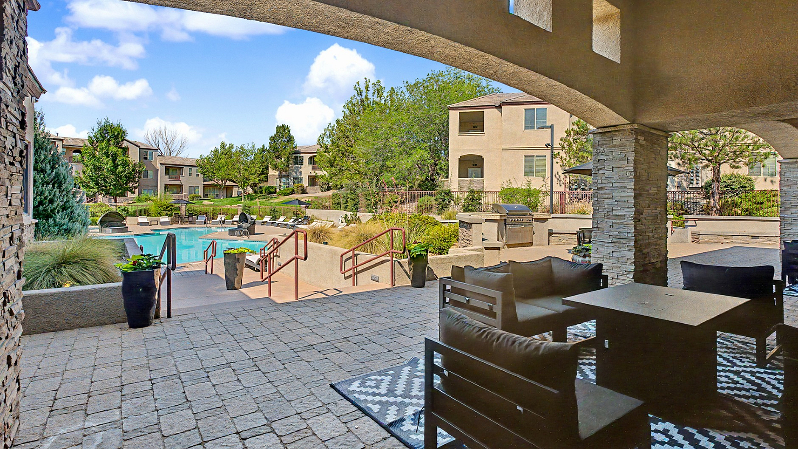 Inviting apartment complex patio with plush seating, cozy outdoor dining area, and a view of a luxurious pool surrounded by well-maintained gardens and modern buildings.