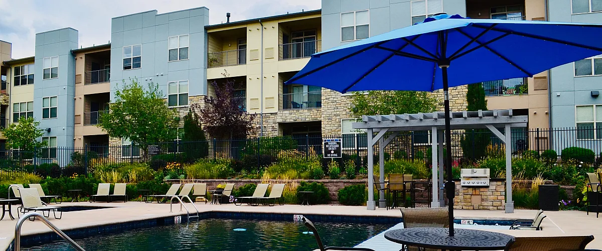 Chic apartment complex with a refreshing pool, stylish lounge chairs, and vibrant landscaping under a spacious blue umbrella.