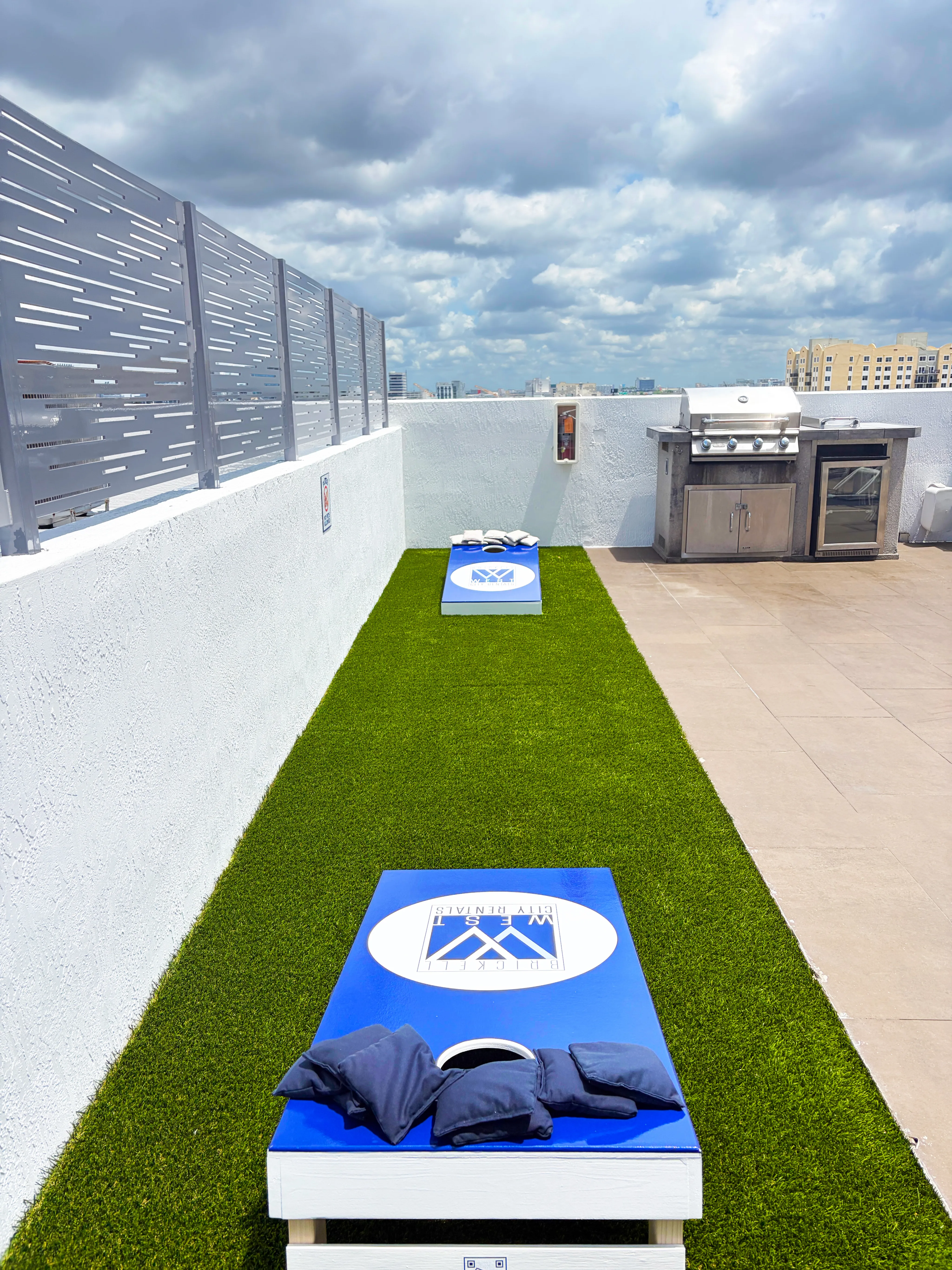 Vibrant rooftop area featuring green turf, a cornhole game set, and a modern outdoor grill with stunning cityscape views.