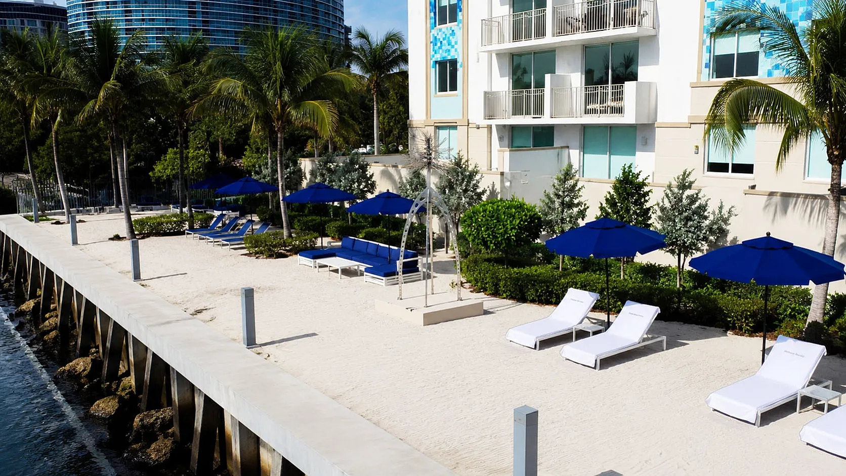 Luxury waterfront apartment exterior with chic beachside lounge area, featuring sunbeds, blue parasols, and vibrant greenery.