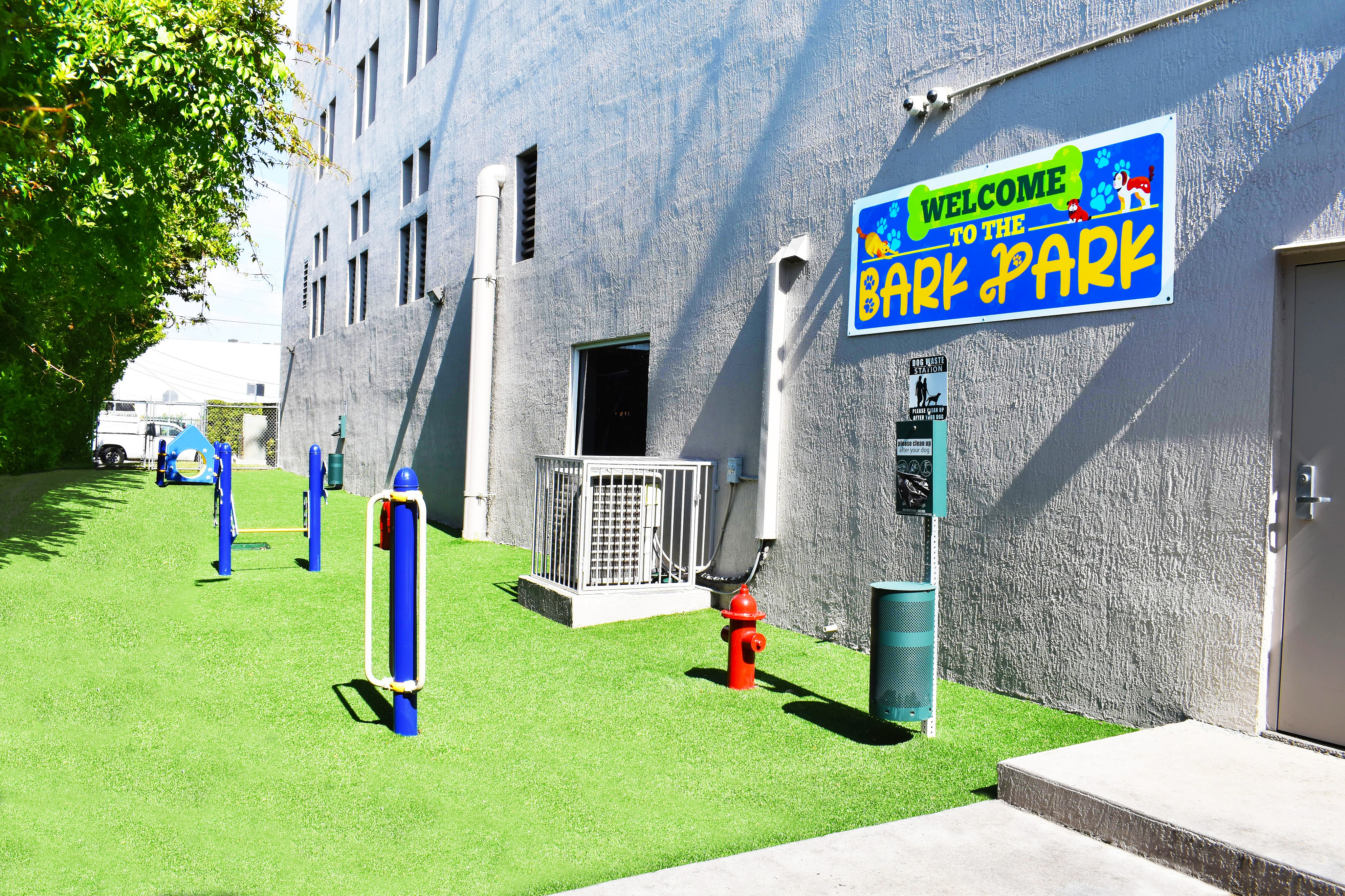 Vibrant pet-friendly apartment amenity featuring a colorful \'Welcome to the Bark Park\' sign, agility equipment, and lush green turf.