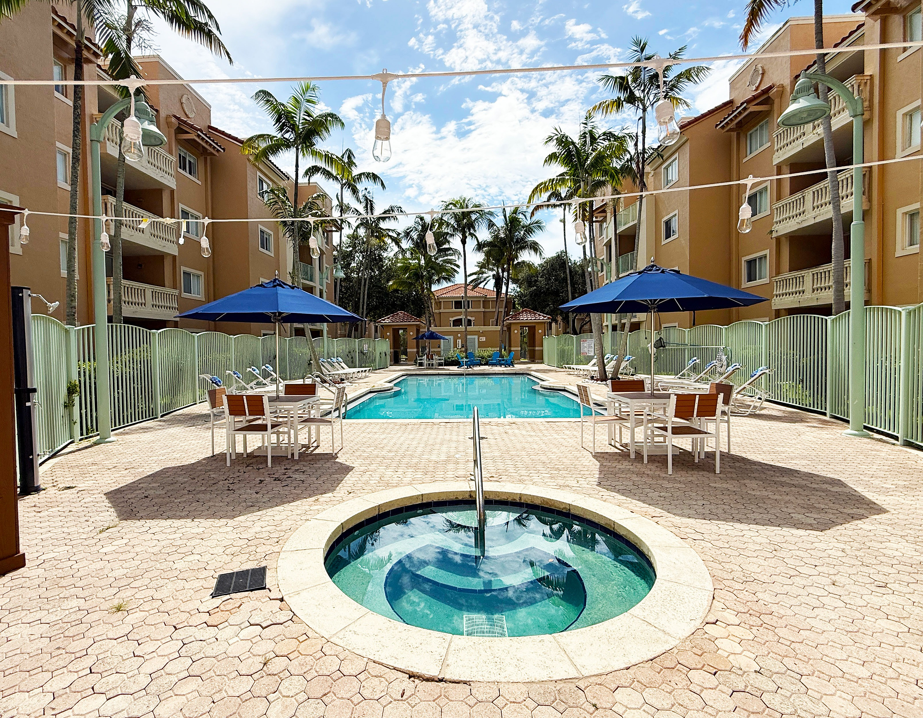 Luxurious outdoor pool area with hot tub, surrounded by stylish loungers, umbrellas, and lush palm trees, set in a chic apartment complex.