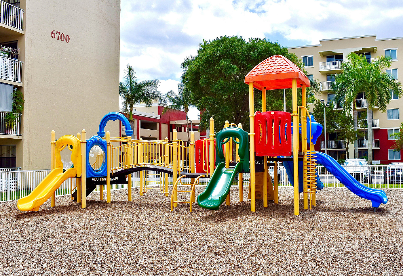Vibrant playground with colorful slides and climbing structures, set against a backdrop of modern apartment buildings and lush palm trees.
