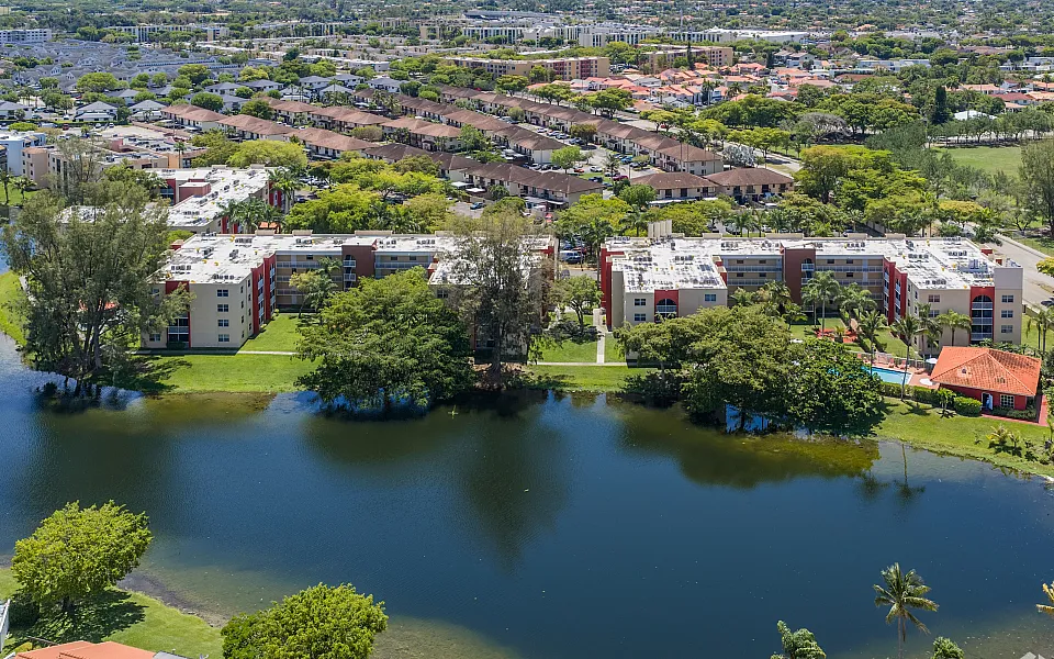 Scenic aerial view of a serene apartment complex surrounded by lush greenery, with a calming lake and vibrant neighborhood backdrop.
