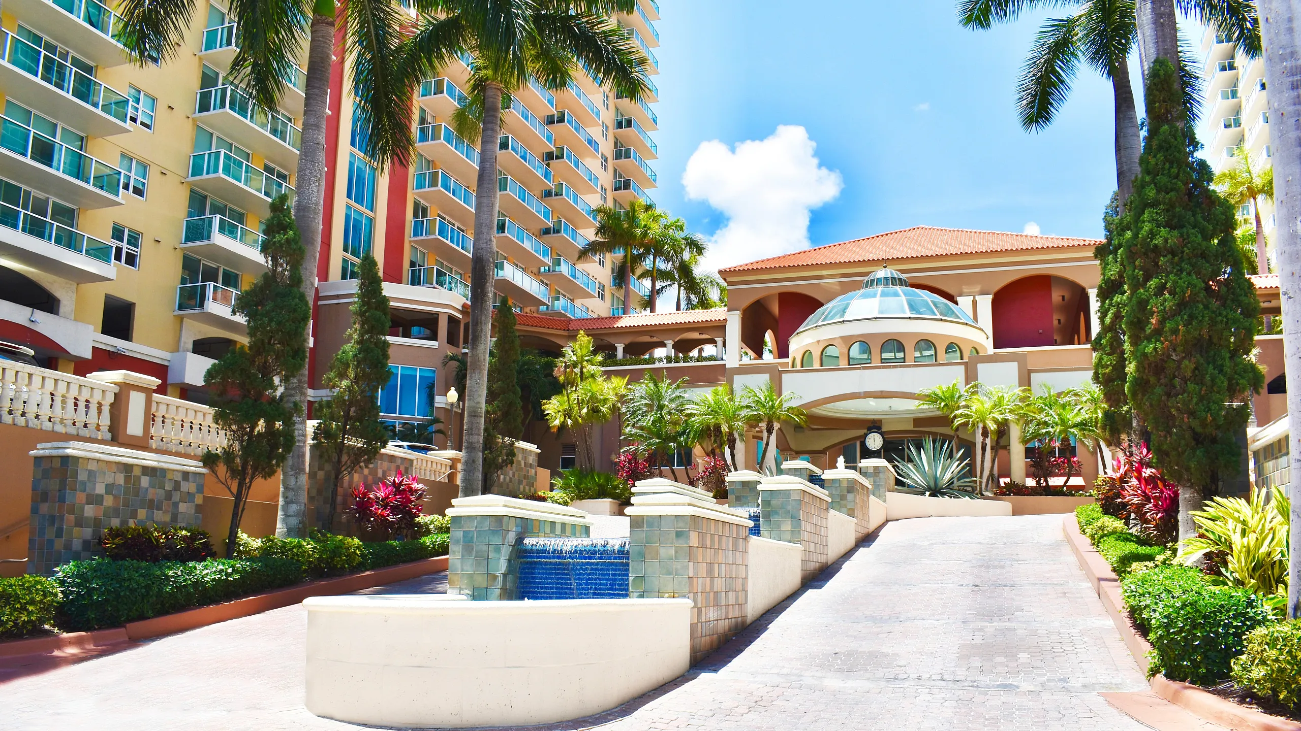 Elegant apartment complex entrance with palm trees, vibrant landscaping, and a luxurious fountain under a bright blue sky.