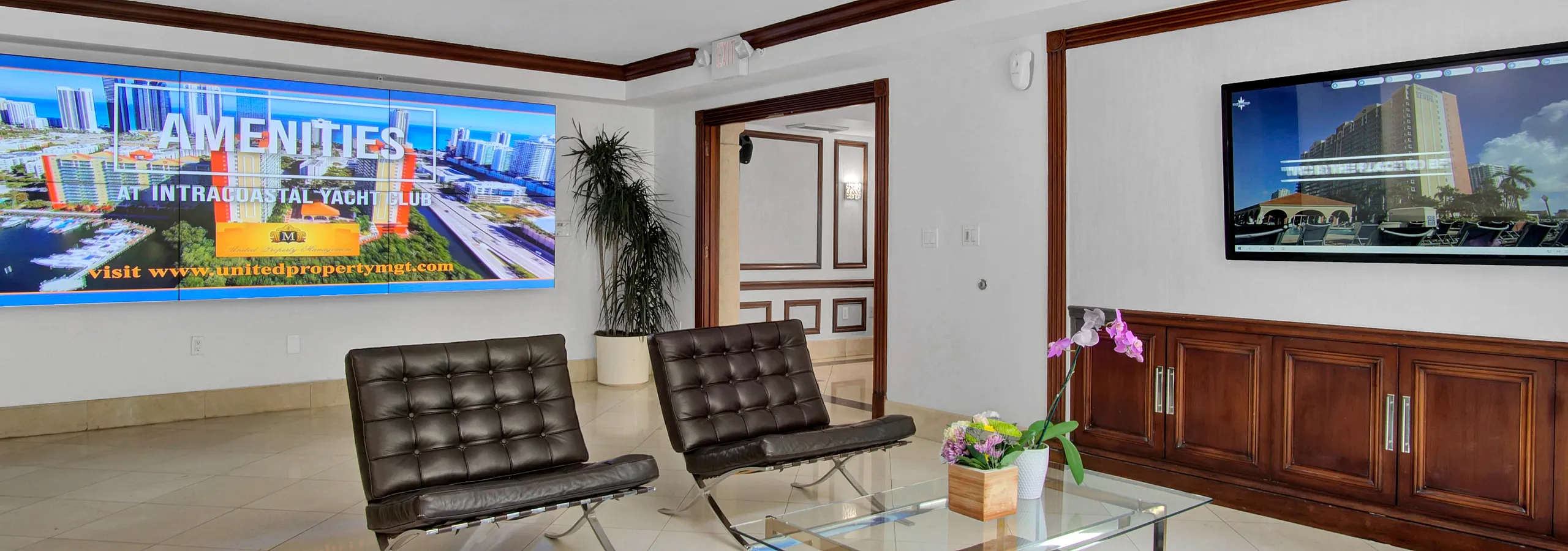 Chic and modern lounge area featuring elegant leather chairs, a glass coffee table, and bright screens showcasing amenities at Intracoastal Yacht Club, complemented by a striking ceiling light and vibrant decor.