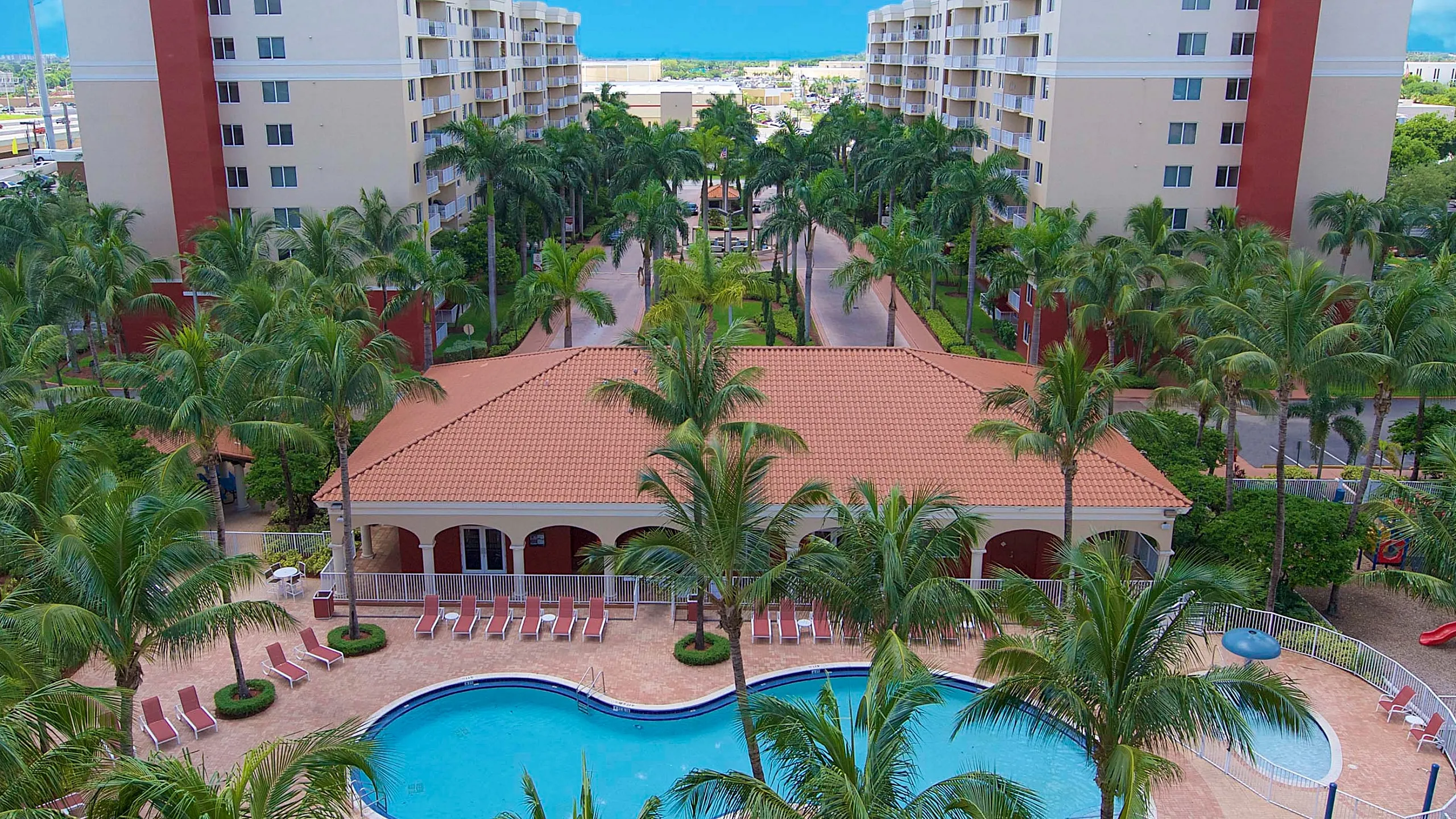 Luxurious apartment complex featuring stunning resort-style pool, lush palm landscaping, and elegant architecture under a vibrant blue sky.