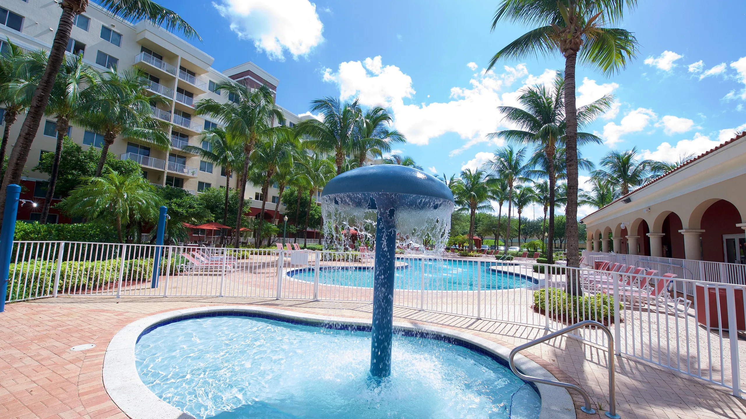Elegant apartment complex with a resort-style pool area, lush palm trees, and a cascading water feature, perfect for relaxation and leisure.