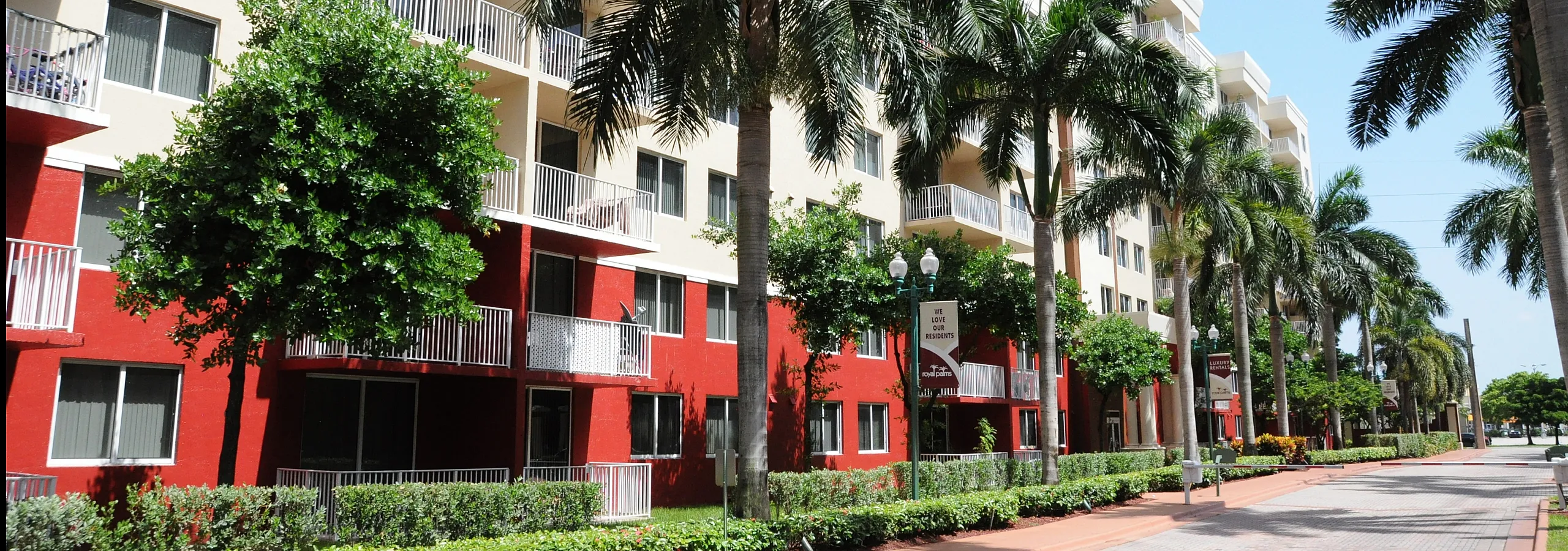 Elegant multi-story apartment building with vibrant red and cream facade, framed by lush palm trees and a welcoming pathway.