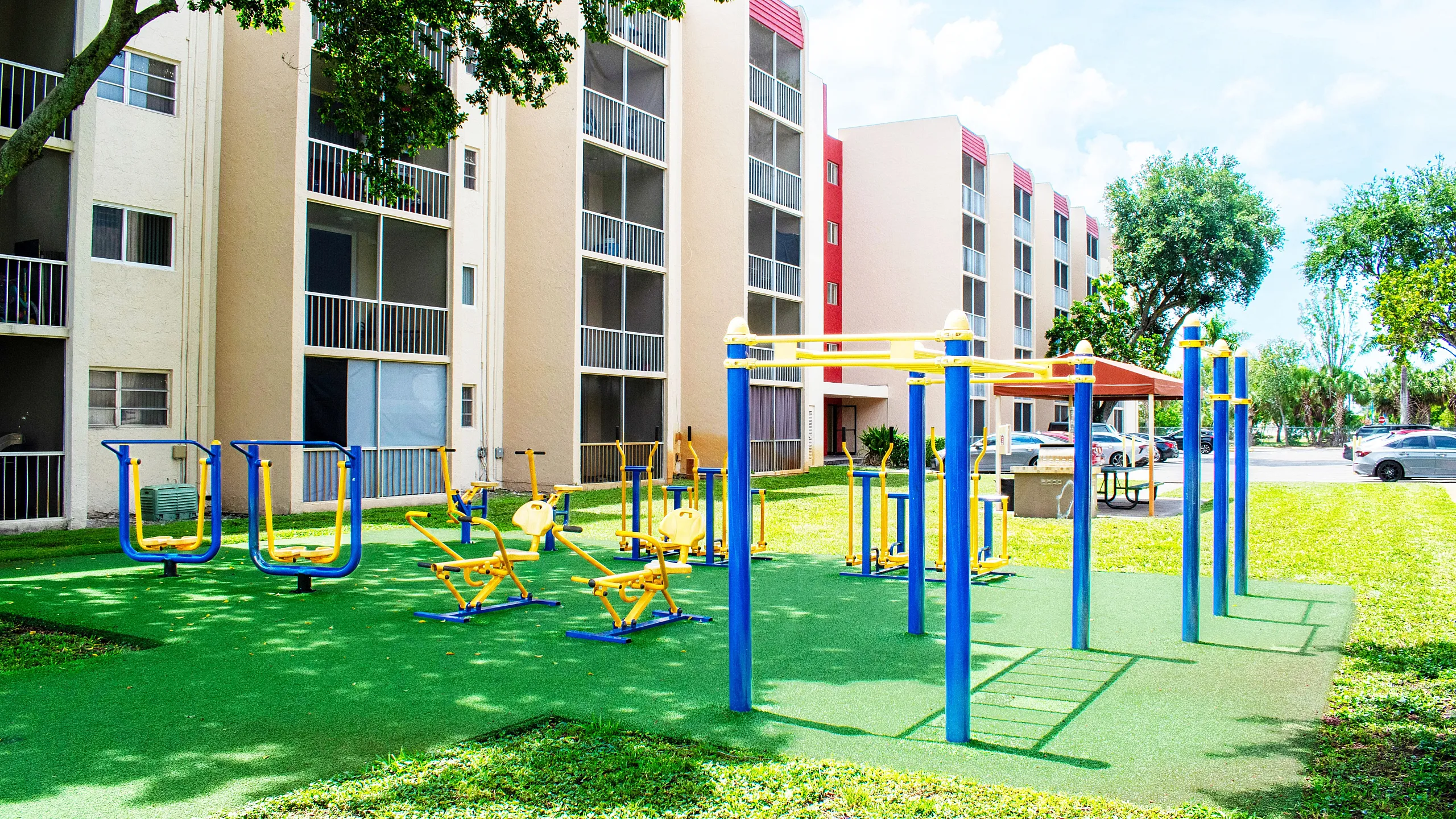 Vibrant apartment complex with an outdoor fitness area featuring colorful exercise equipment surrounded by lush greenery.