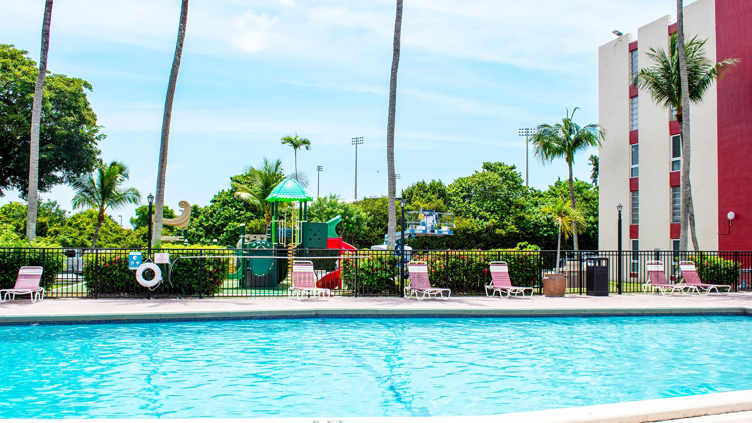 Vibrant apartment community pool area with palm trees, lounge chairs, and a colorful playground under sunny skies.