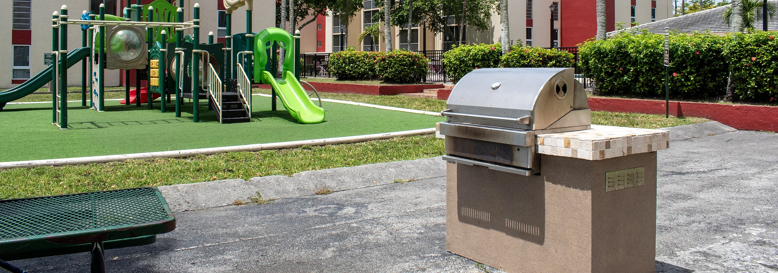 Vibrant apartment community with a modern playground, lush palm trees, and a handy outdoor grill area, perfect for family gatherings.