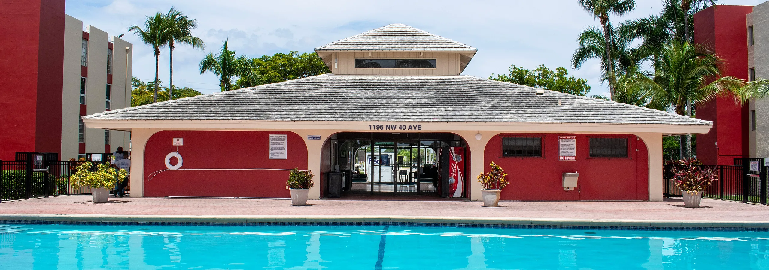 Inviting apartment complex with a sparkling swimming pool, flanked by vibrant palm trees and a stylish red and cream clubhouse, perfect for relaxation and socializing.