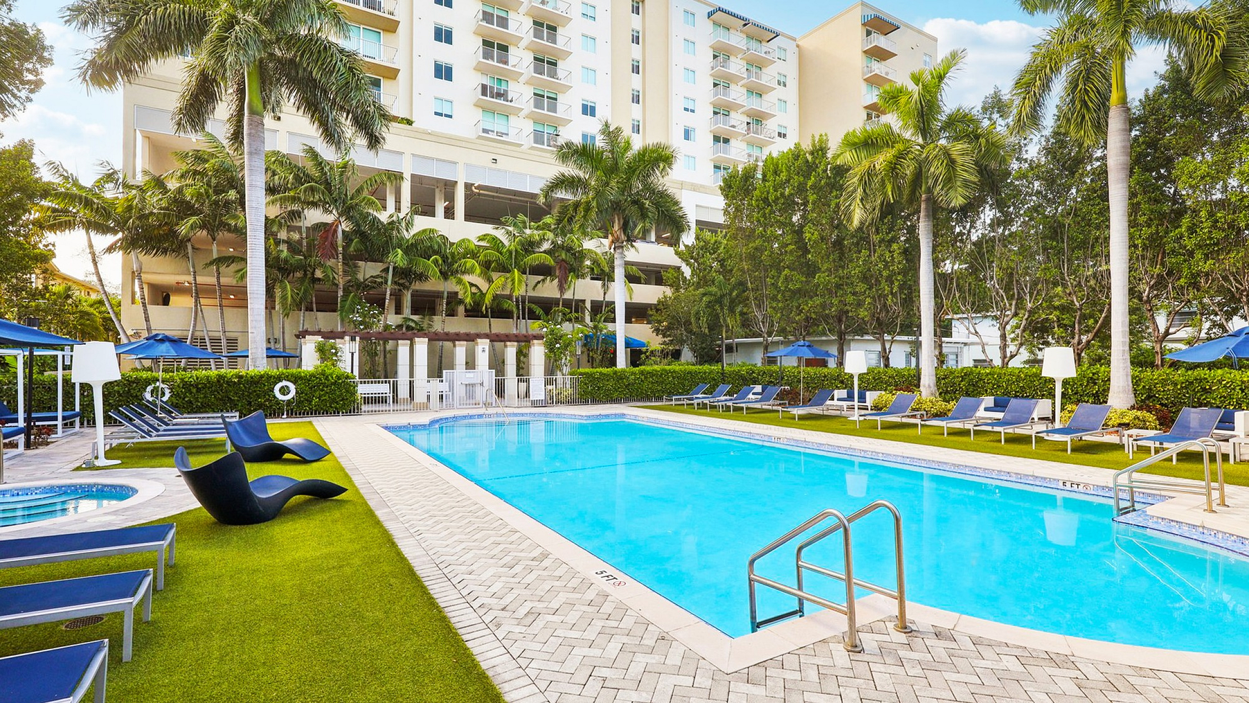 Luxurious apartment complex with tropical vibes featuring a pristine swimming pool surrounded by lush palm trees and elegant lounge chairs.