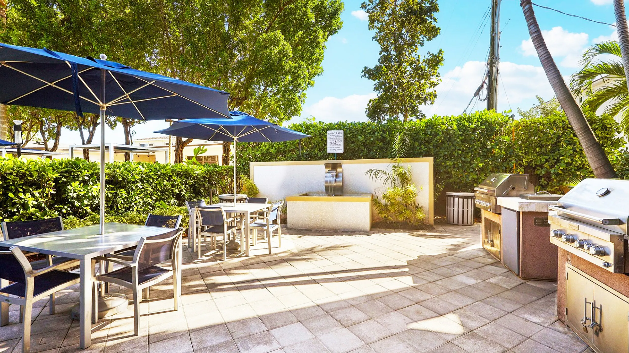 Elegant outdoor patio with modern dining area, shaded by blue umbrellas, featuring lush greenery and high-end BBQ grills.