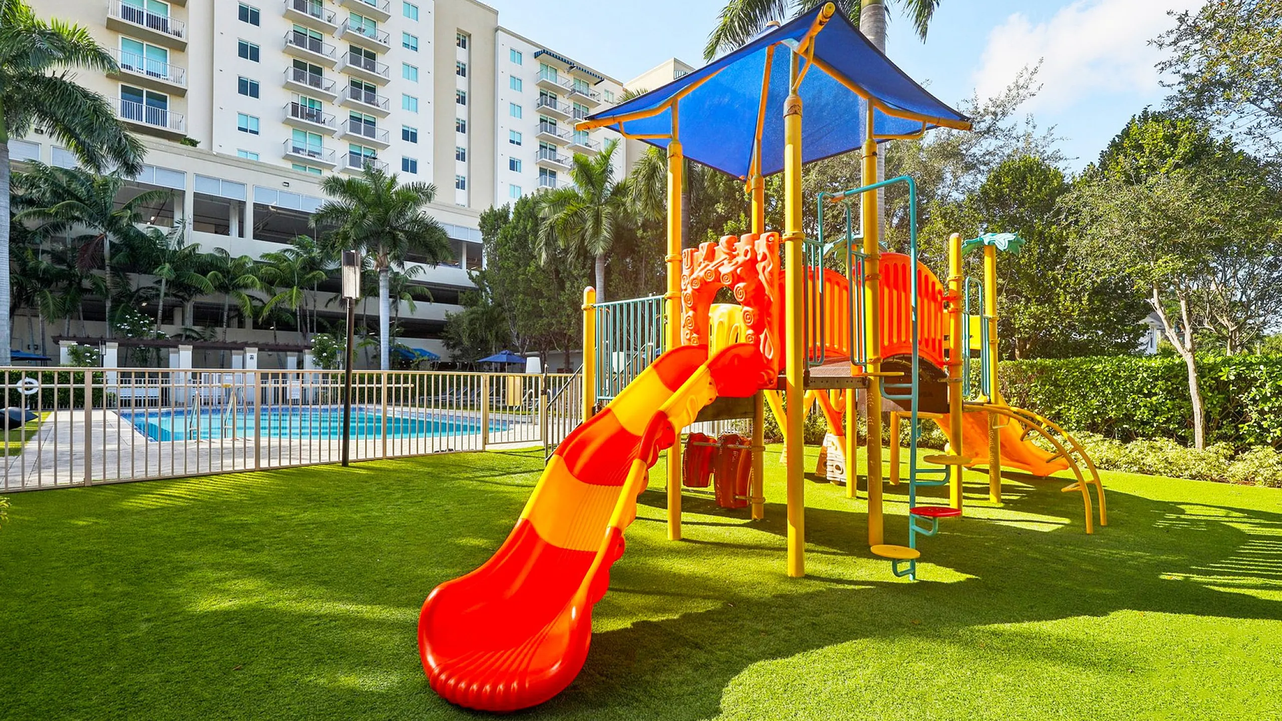 Vibrant apartment community featuring a colorful children's playground and sparkling swimming pool, surrounded by lush greenery and modern high-rise buildings.