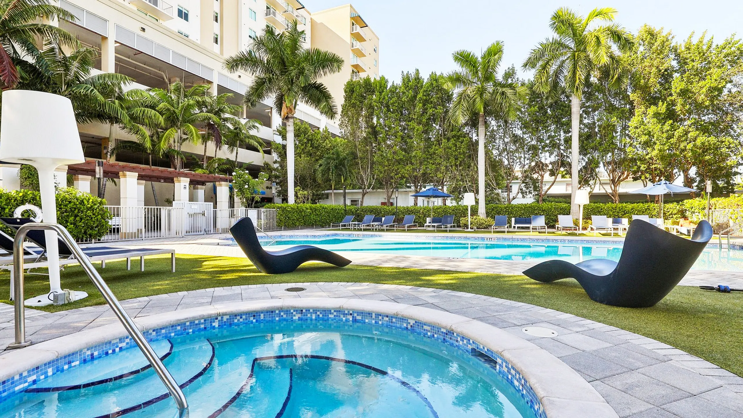 Luxurious apartment complex featuring a sparkling pool area surrounded by lush palm trees, modern lounge chairs, and immaculate landscaping.