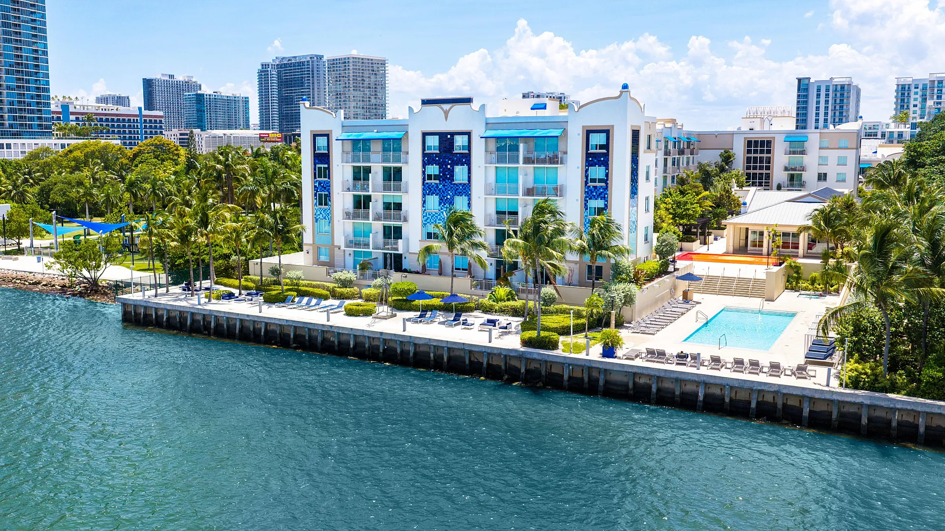 Chic waterfront apartments with vibrant blue accents, surrounded by lush palm trees and featuring a luxurious swimming pool beside the serene waterway.