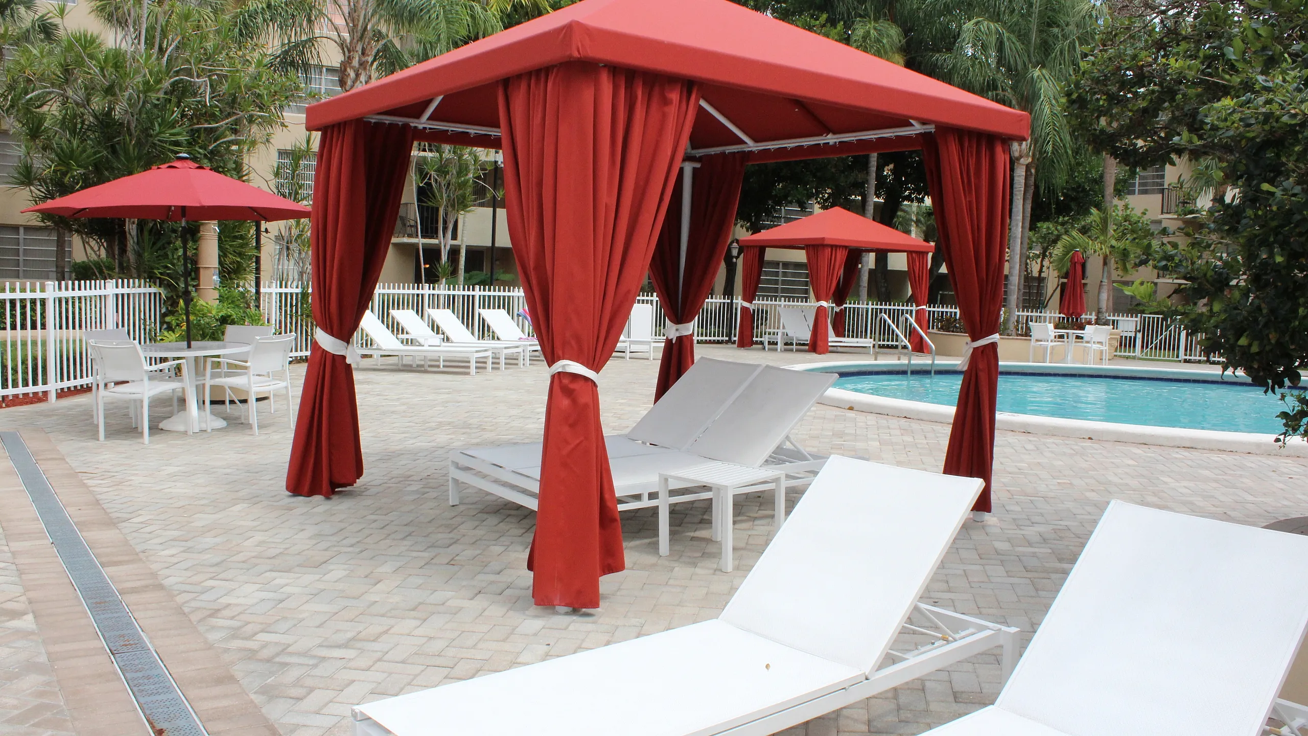 Luxurious poolside cabanas with chic red canopies and stylish white lounge chairs create a relaxing oasis surrounded by lush greenery.