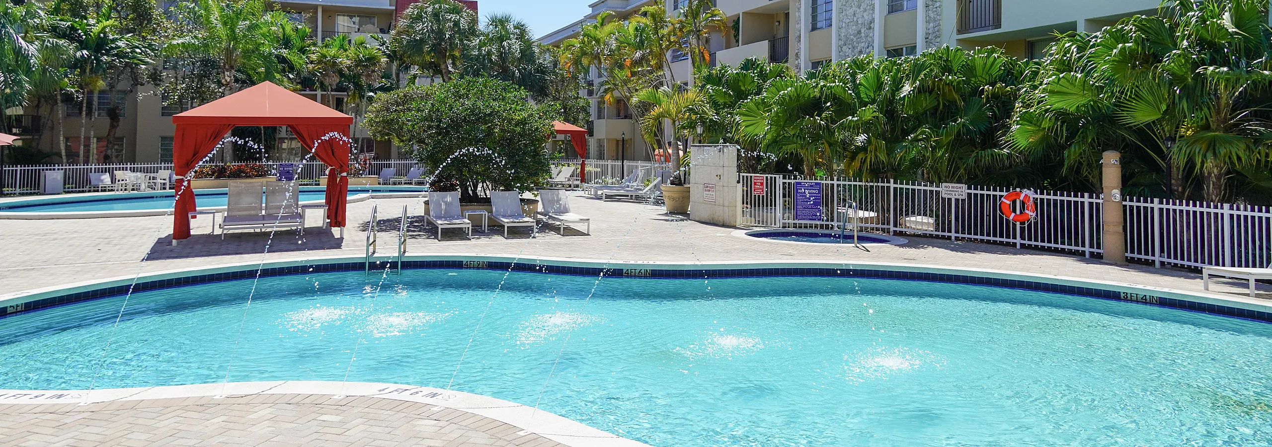 Charming apartment complex with modern amenities including a sparkling swimming pool, lush landscaping, and inviting lounge areas under red canopies, perfect for relaxation.