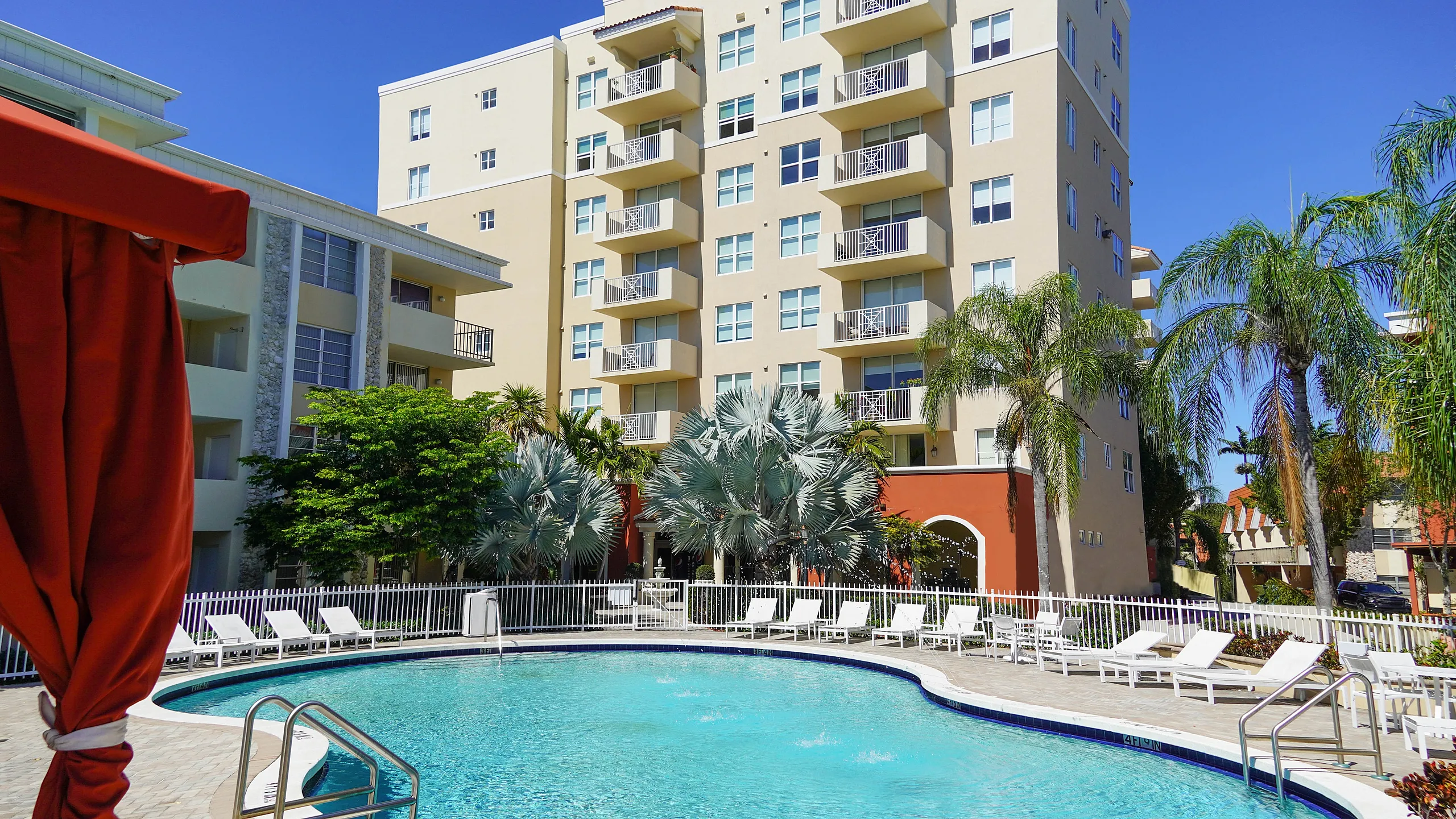 Luxurious apartment complex with a sparkling pool, surrounded by lush palm trees and modern architecture under a clear blue sky.