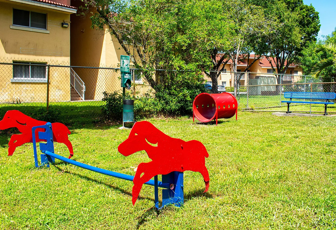 Inviting outdoor dog play area featuring vibrant red agility equipment, green grass, and a nearby park bench under shaded trees.