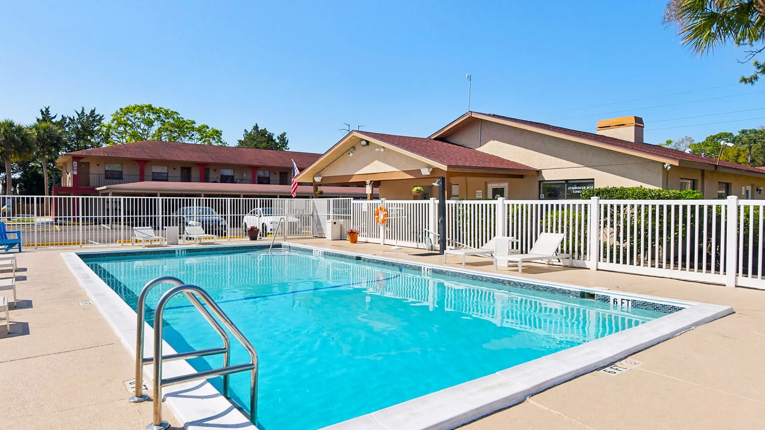 Inviting outdoor pool area with modern loungers, surrounded by a secure white fence, set against a charming apartment backdrop, ideal for a relaxing day under the sun.