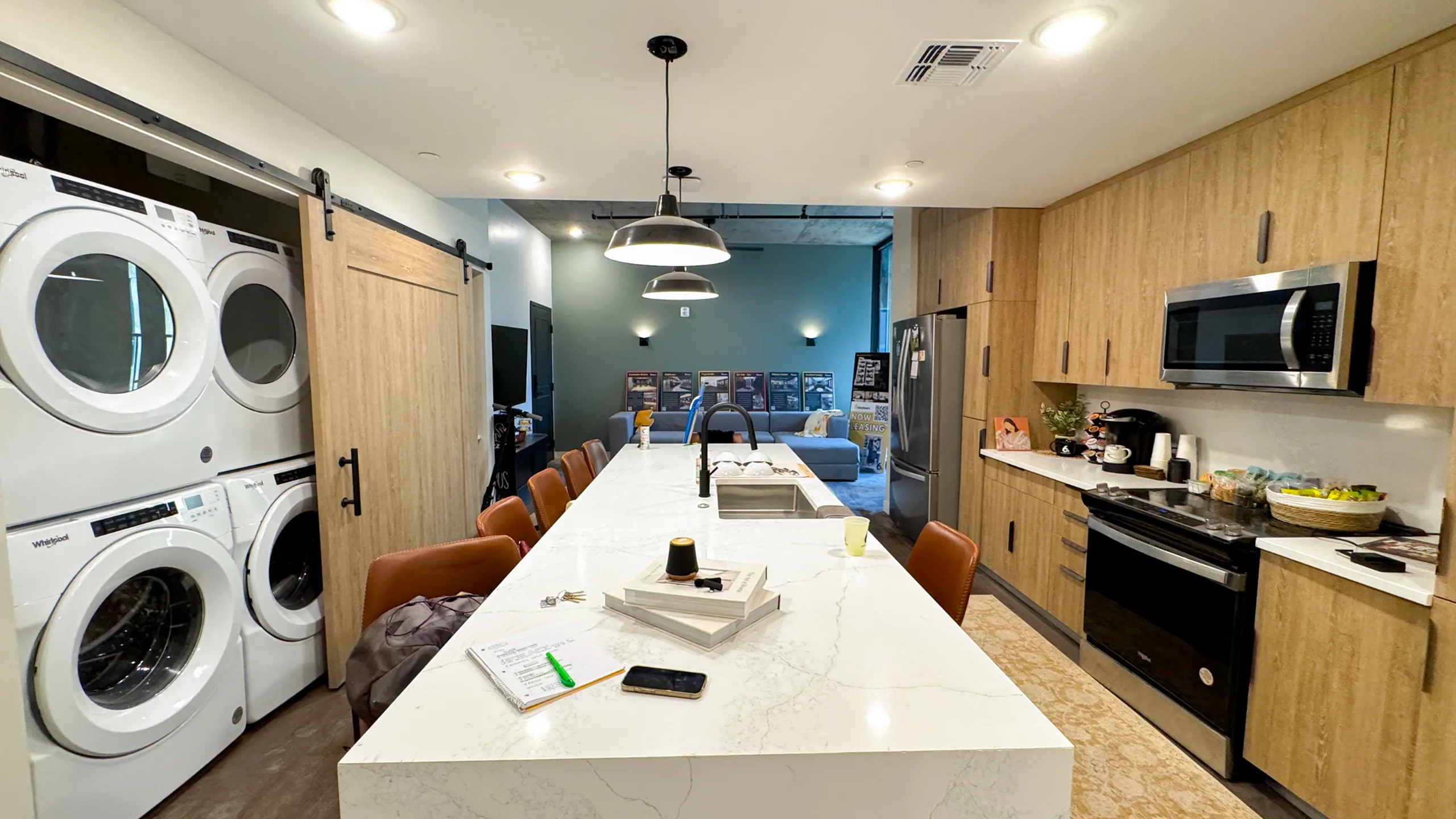 A modern communal kitchen and living area with a long white marble island, tan leather bar stools, stacked Whirlpool washers and dryers behind a wooden sliding door, stainless steel appliances, and light wood cabinetry. A cozy living room w