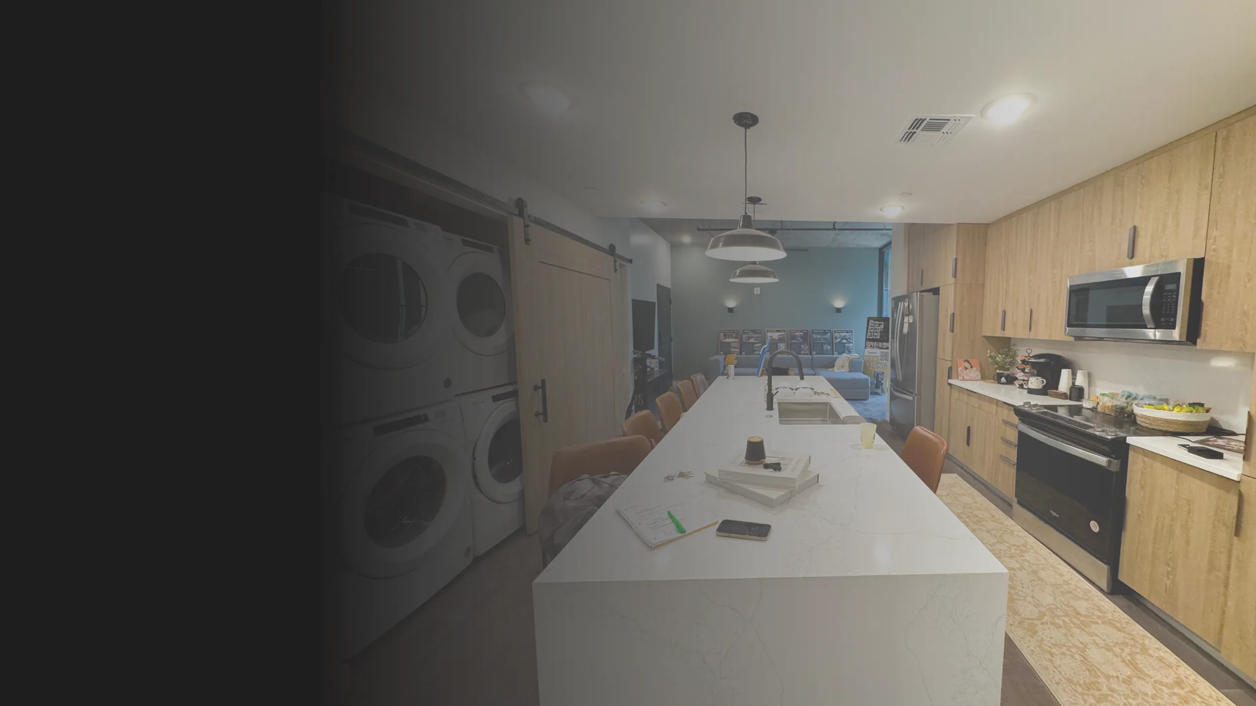 Modern communal kitchen with a long marble island, bar stools, stacked washer/dryer units, wood cabinetry, and stainless steel appliances.