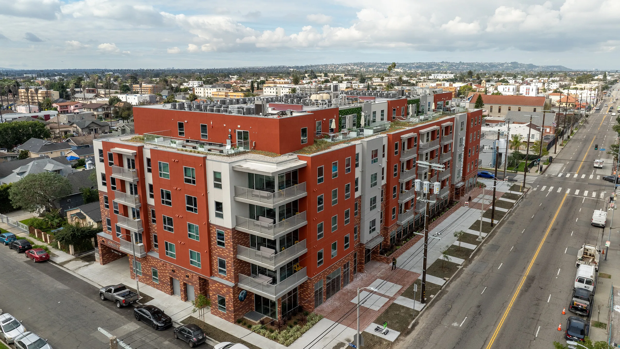 Aerial view of a modern multi-story apartment complex with a red and white fa&ccedil;ade, rooftop terraces, and private balconies. The building is situated on a corner lot with wide sidewalks and landscaped streets, surrounded by a mix of r