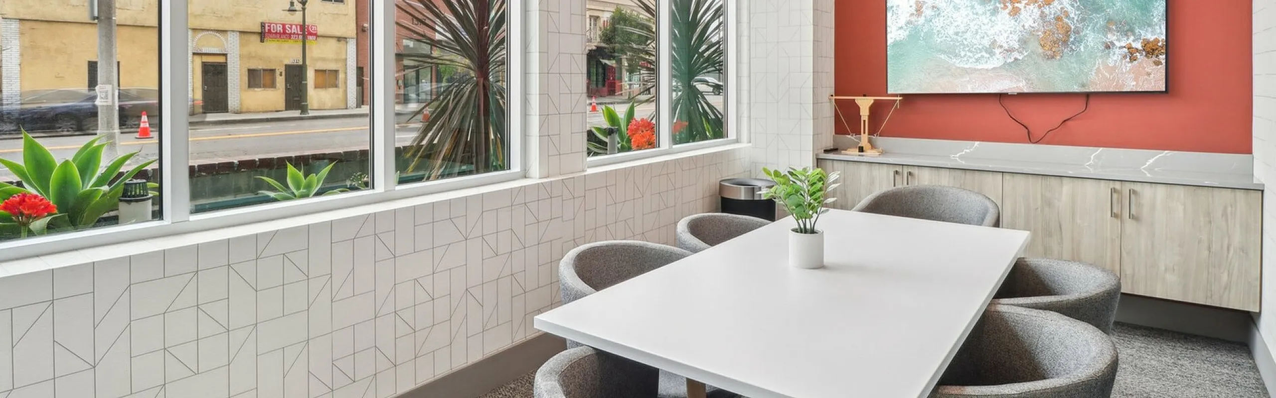 A bright and modern meeting room with floor-to-ceiling corner windows offering views of an urban street. The space features a sleek white table surrounded by gray upholstered swivel chairs. A potted plant sits on the table, and a vibrant re