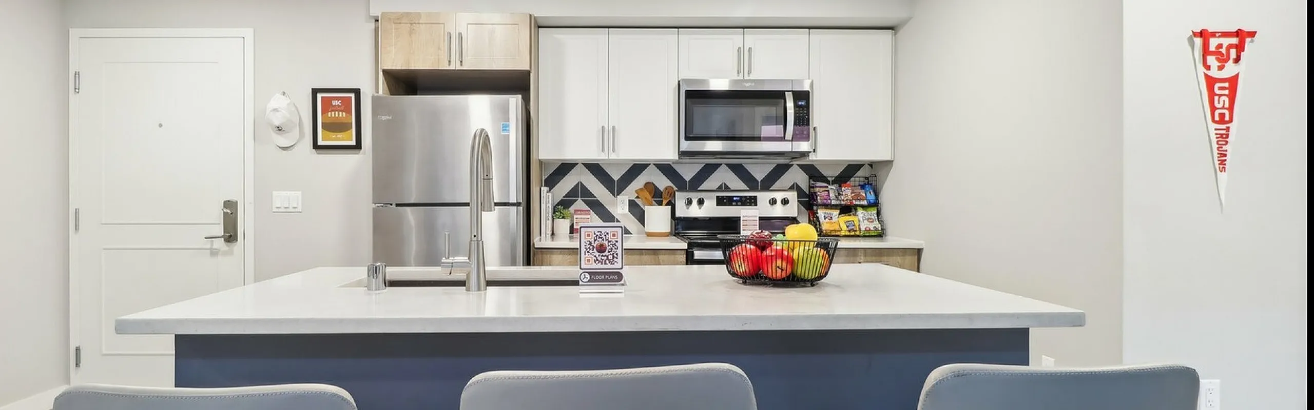 A contemporary kitchen with a central island featuring a white countertop, integrated sink, and three gray barstools. The kitchen is equipped with stainless steel appliances, including a refrigerator, oven, and microwave, and has a bold che