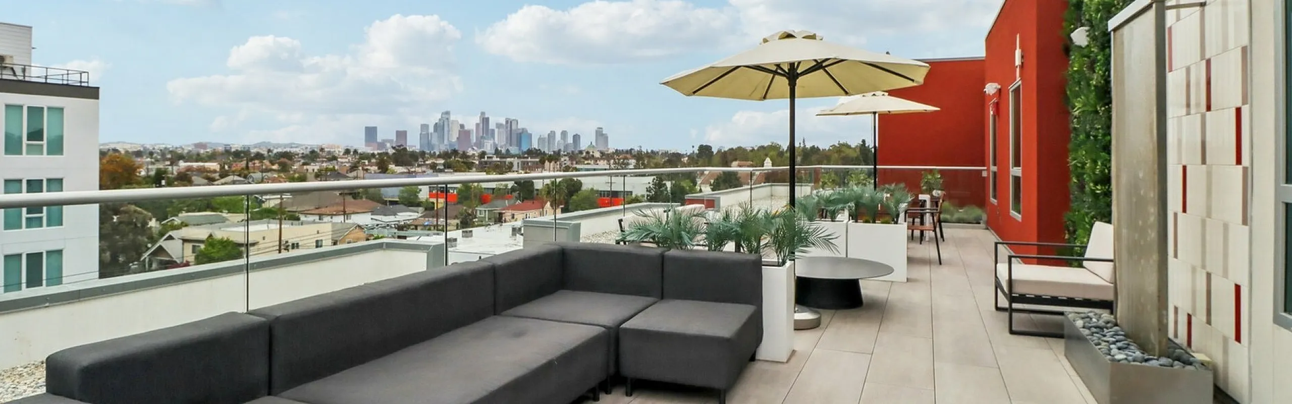 A spacious rooftop lounge with a panoramic view of the Los Angeles skyline. The area features a long modular dark gray sofa, modern round coffee tables, patio chairs, and large beige umbrellas for shade. A living green wall adds natural tex