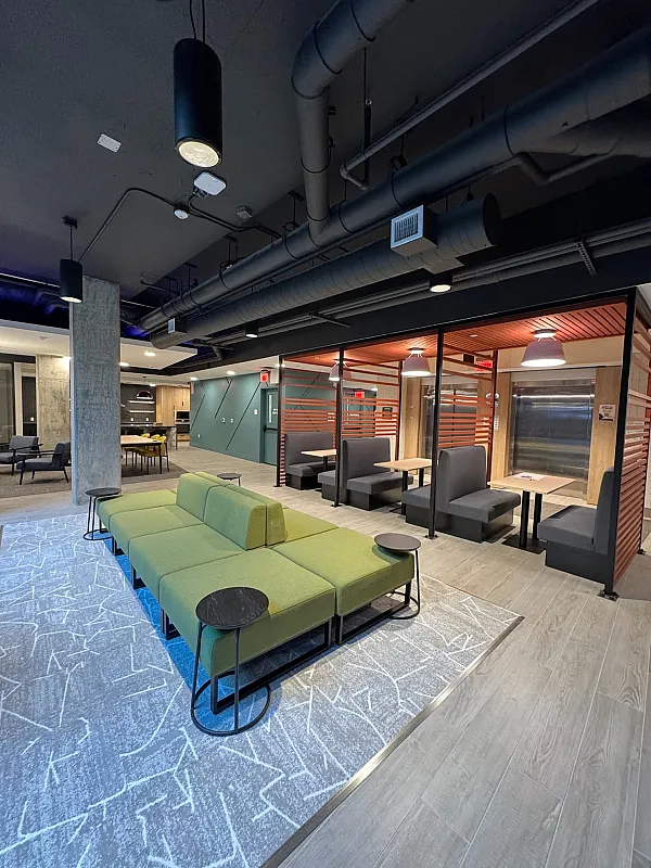 Modern communal lounge area with green modular sofas and small round black side tables on a patterned gray rug. In the background, there are booth seating areas with wooden accents and overhead lighting, as well as an open kitchen space wit