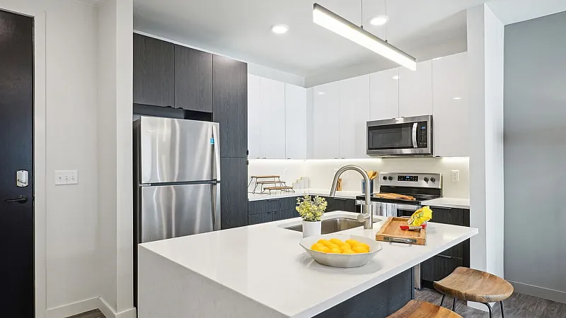 Modern apartment kitchen featuring a sleek island with a bowl of lemons, stainless-steel appliances, two wooden barstools, and bright two-tone cabinetry that highlights the spacious, contemporary layout ideal for everyday living.