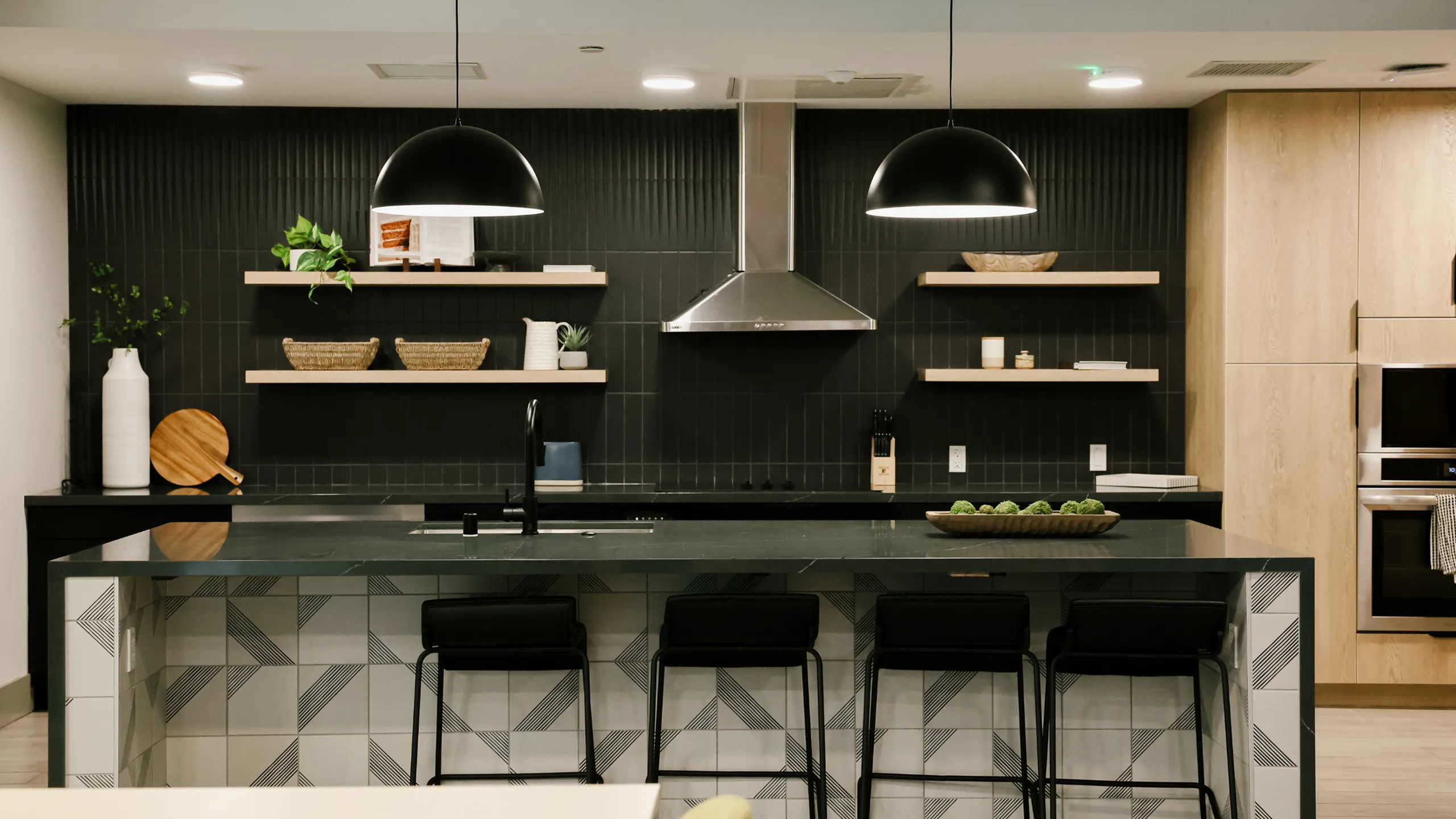 A modern community kitchen at 6Nineteen in San Diego features a sleek black tile backsplash, open wood shelving, and a large island with geometric tile accents ideal for resident gatherings.