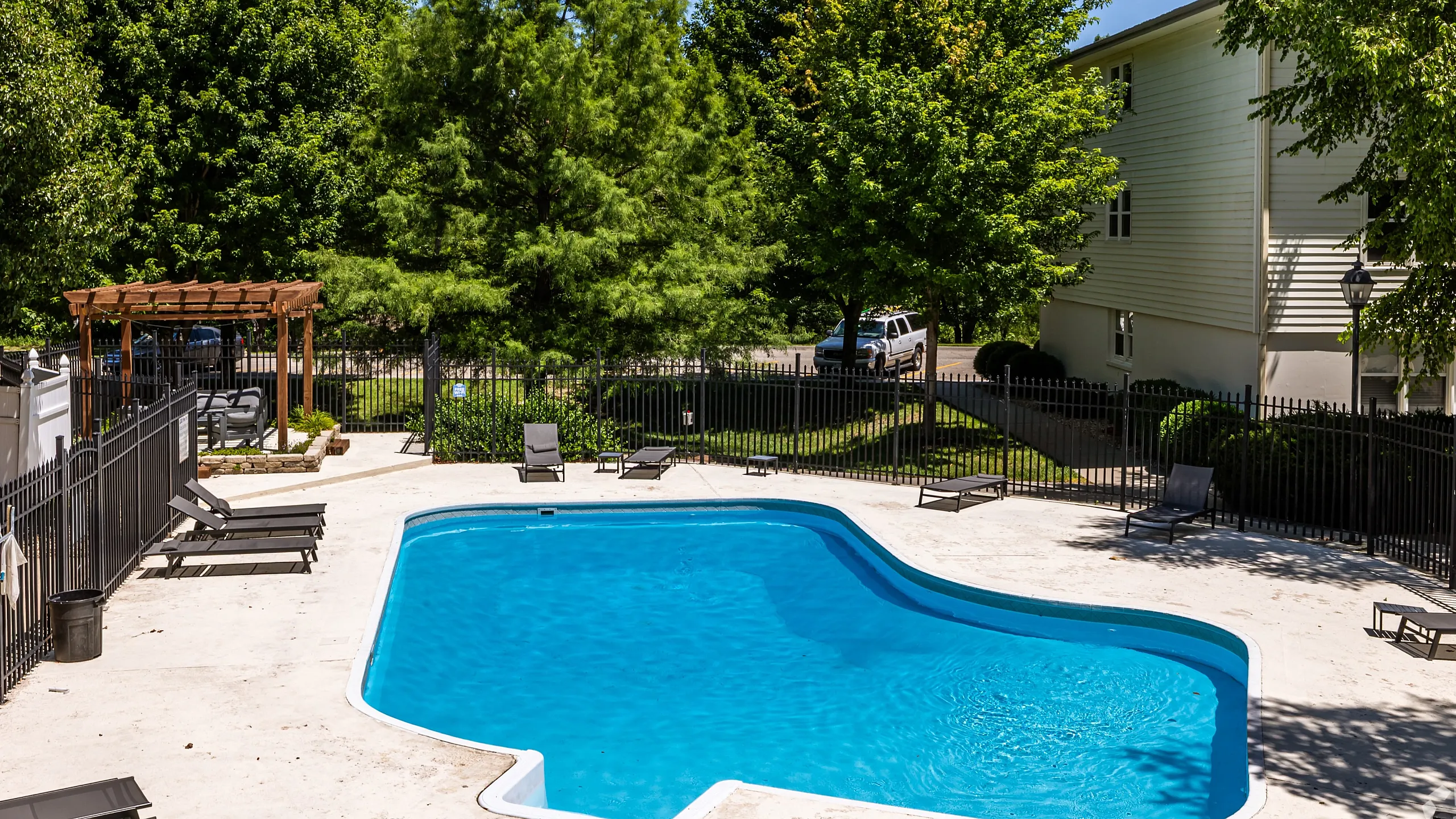 Outdoor swimming pool with lounge chairs and a shaded pergola, set within a fenced apartment community surrounded by mature trees for a relaxing, resort-style amenity.
