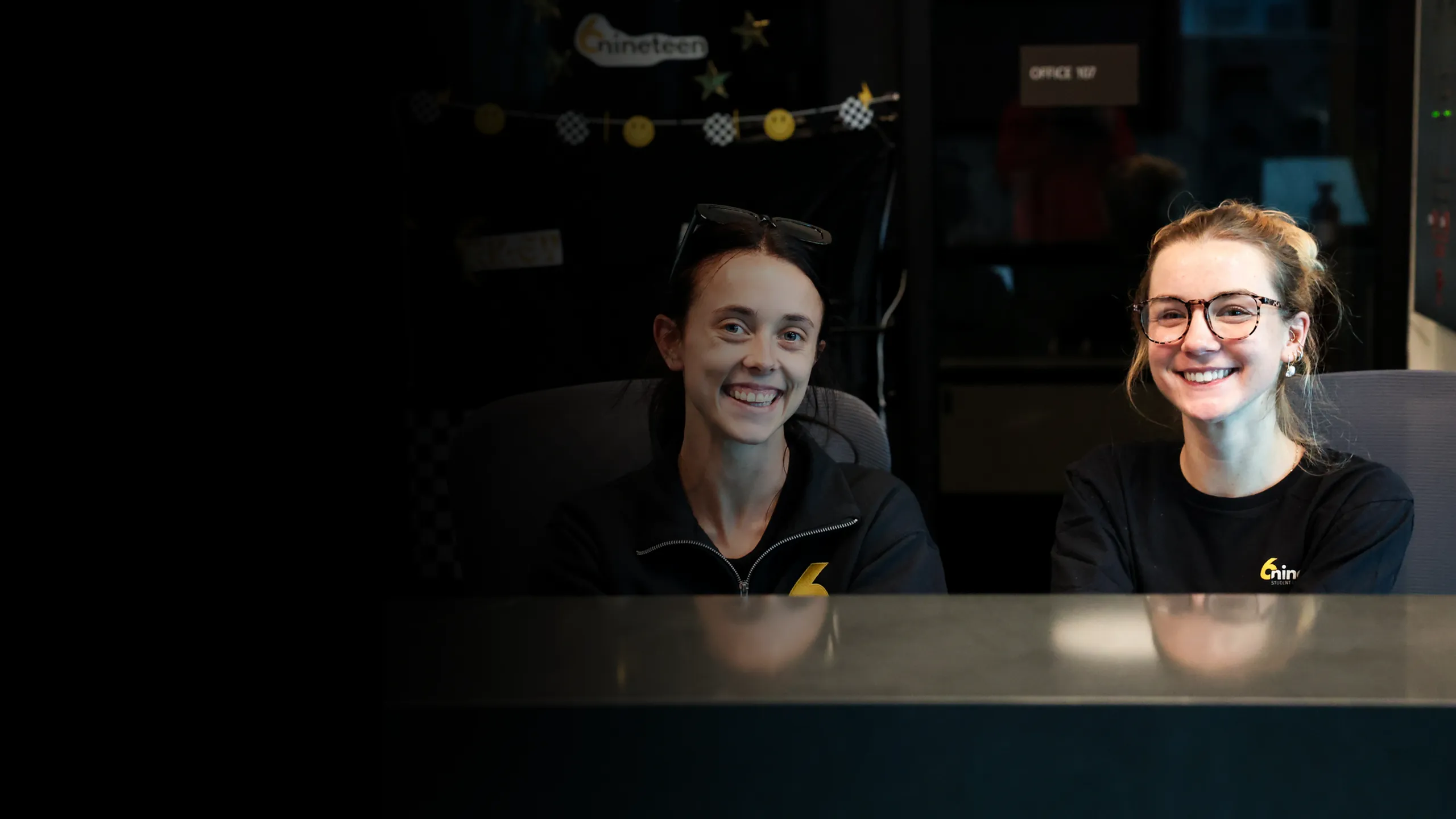 Two smiling front-desk team members sit behind the leasing counter in a modern student-housing office, creating a welcoming first impression for prospects. Their branded apparel and upbeat atmosphere highlight the community&rsquo;s friendly