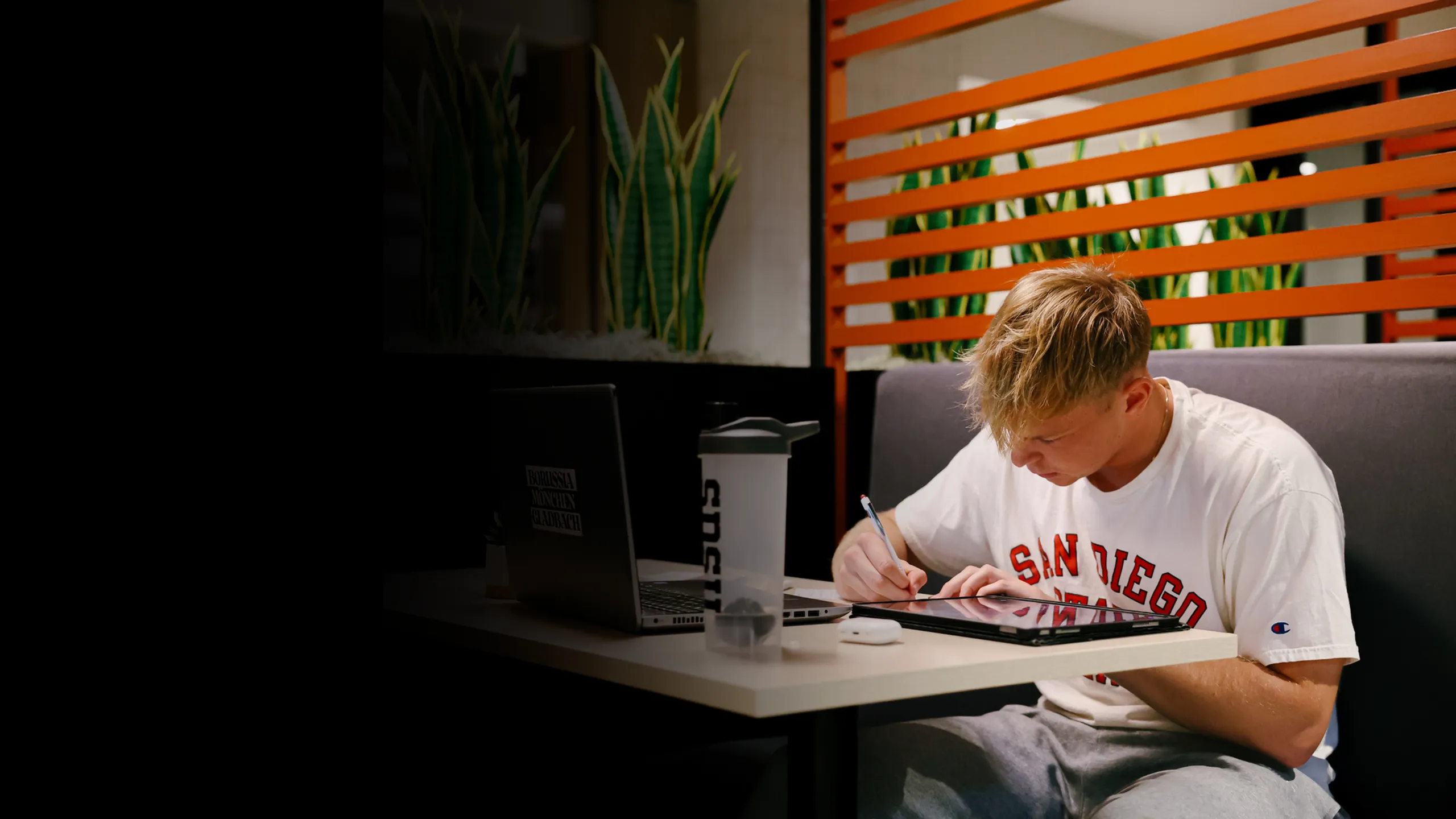 Student studying at a private booth with a laptop and tablet in a modern apartment community study lounge, featuring comfortable seating and greenery for focused, student-friendly living.