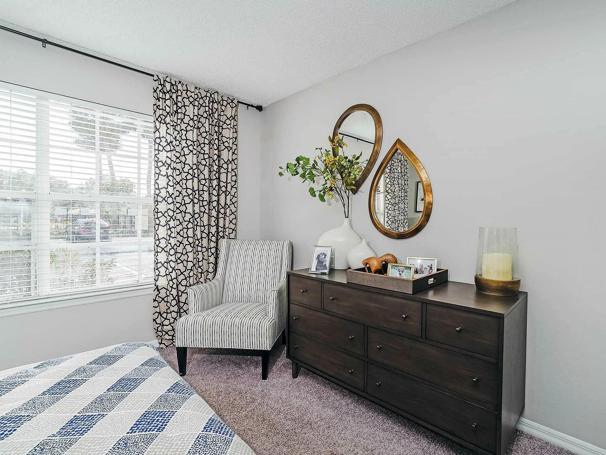 Charming bedroom featuring a stylish dresser with decorative mirrors, a cozy chair, and a large window with patterned curtains.