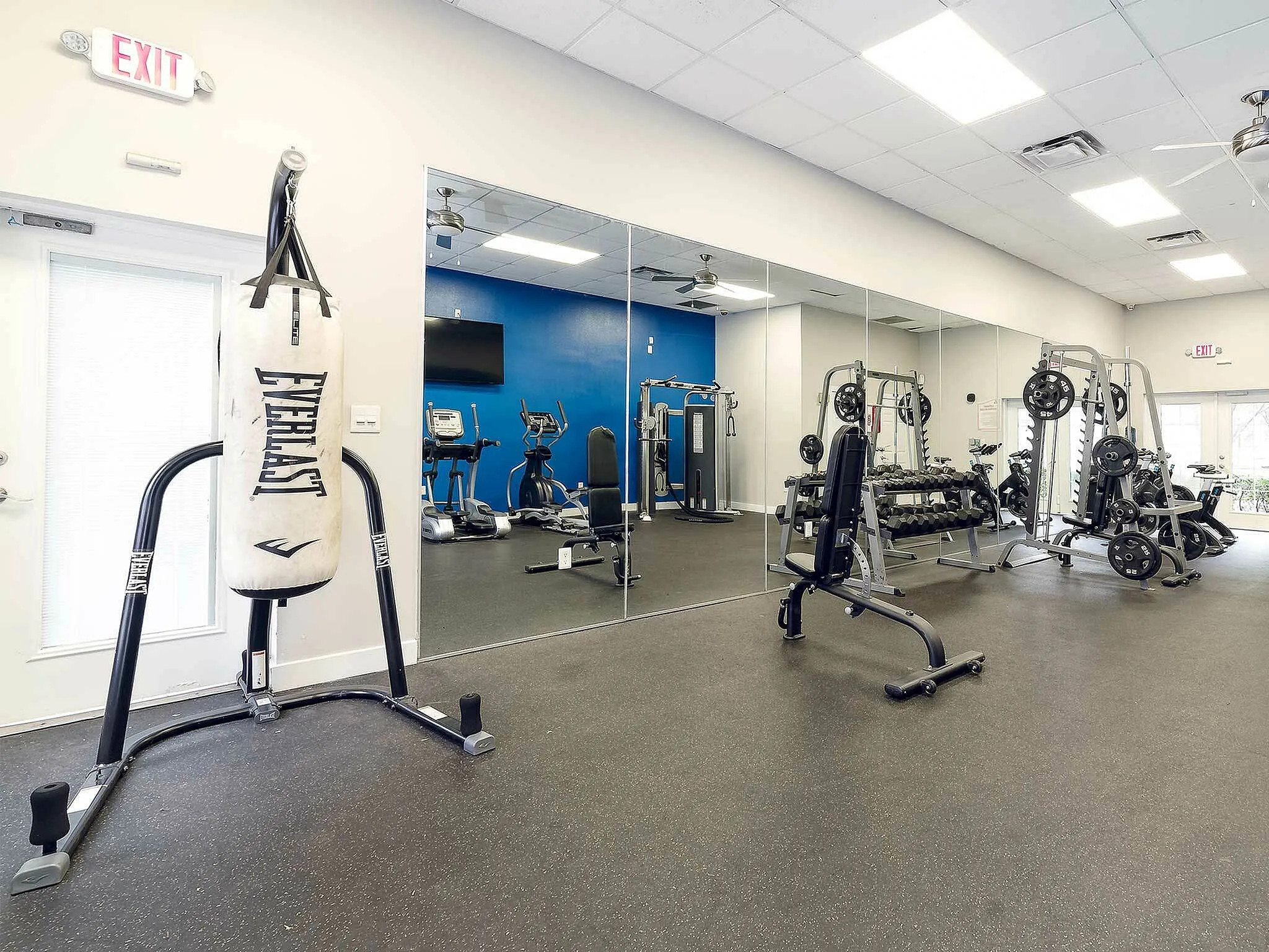 Modern gym room with various exercise equipment including a punching bag, elliptical machines, weight racks, and mirrored walls.