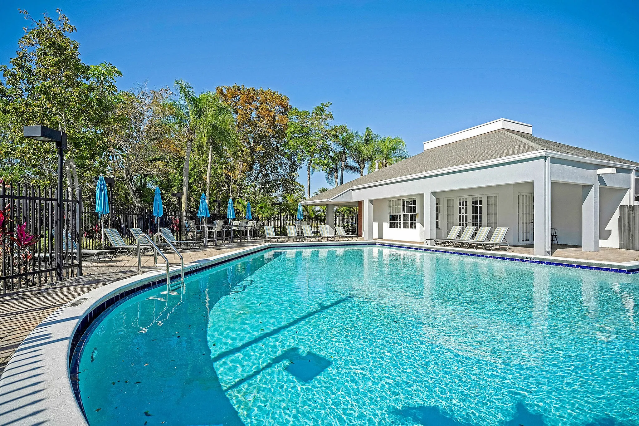 Inviting outdoor pool area with vibrant blue umbrellas and sun loungers, surrounded by lush greenery and a modern clubhouse.