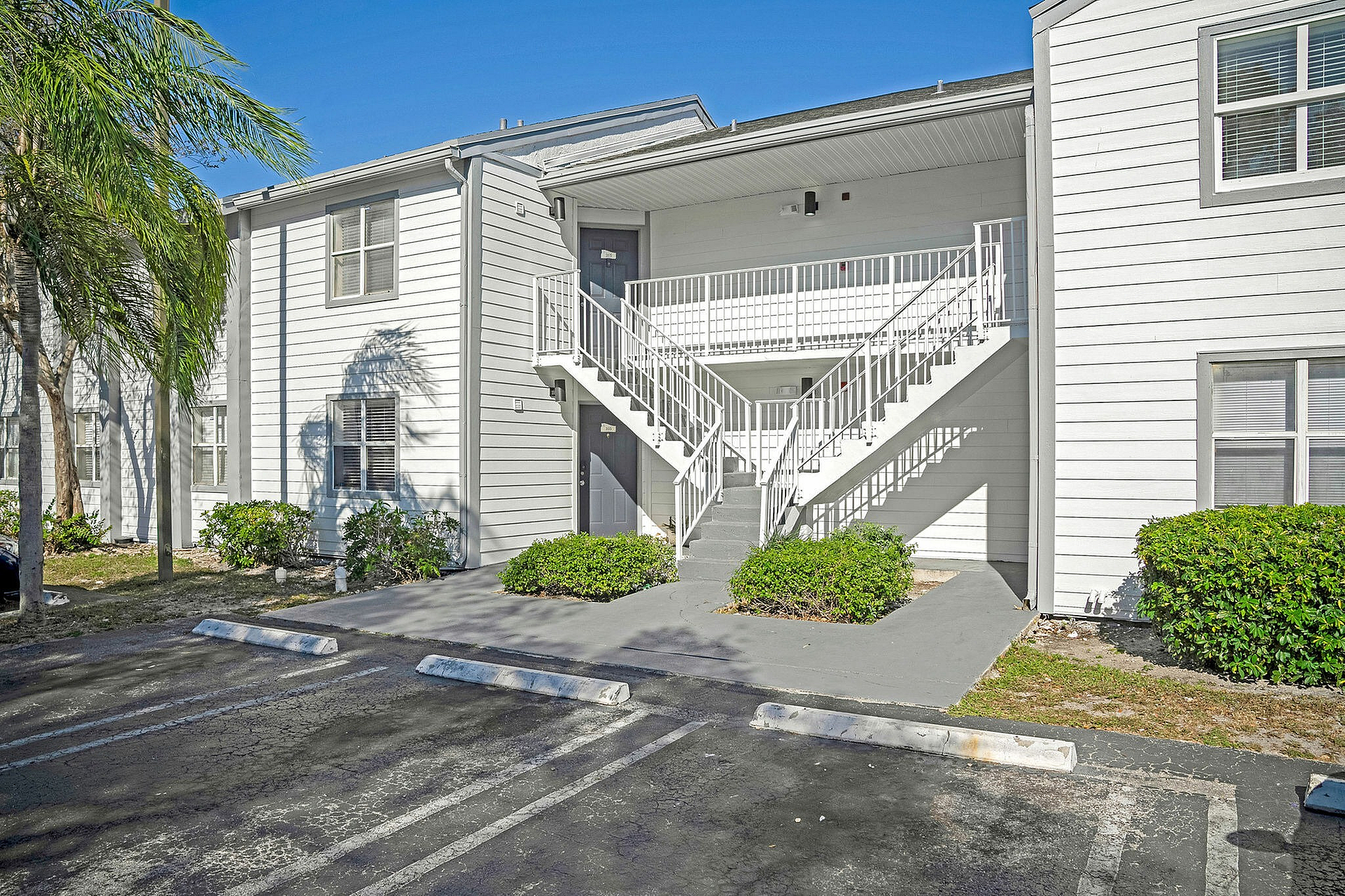 Charming white two-story apartment building with lush green landscaping and modern stairway access, offering serene outdoor parking space.
