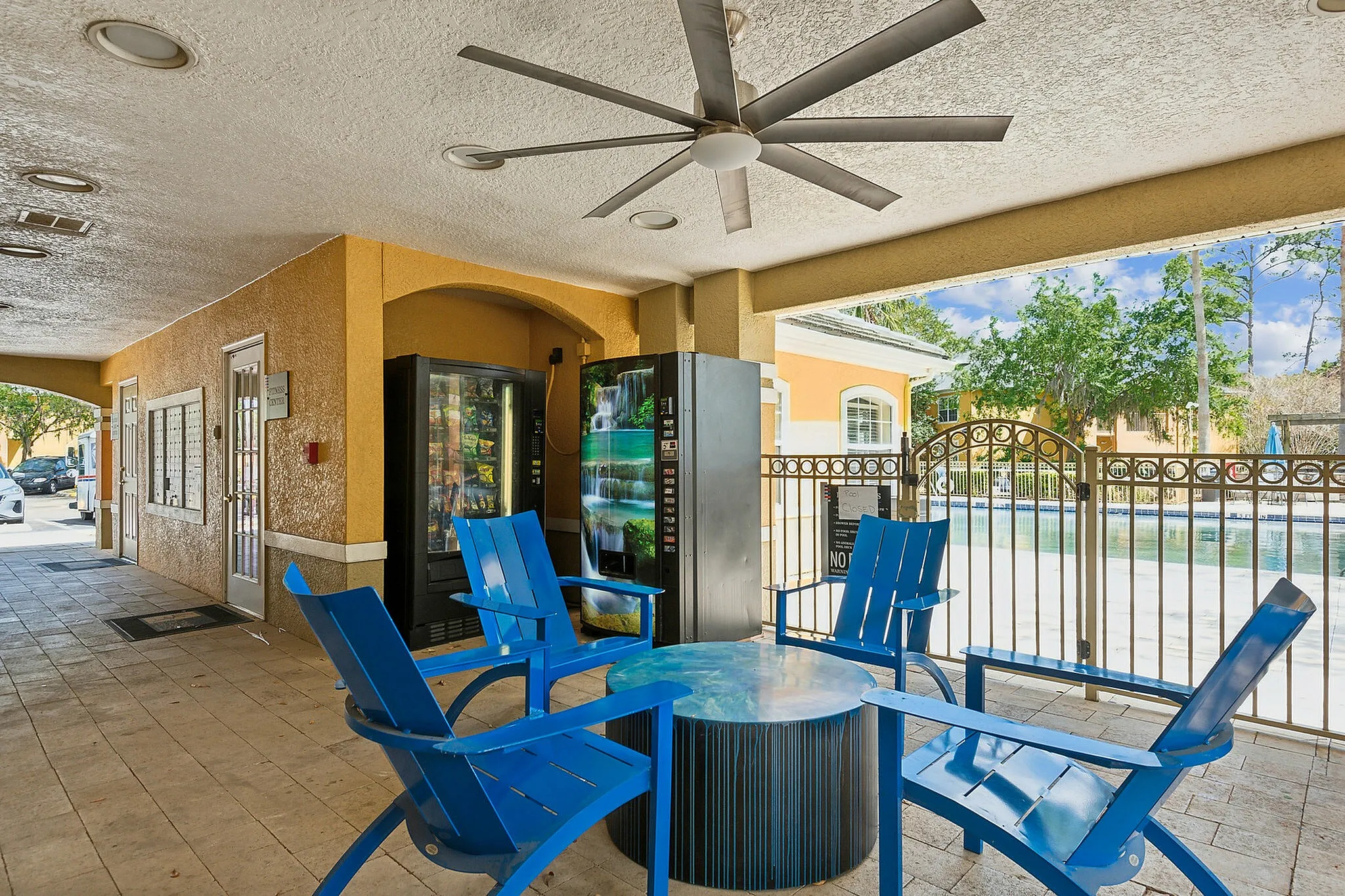 Inviting outdoor lounge area with vibrant blue chairs and a ceiling fan, overlooking a sparkling pool through elegant fencing.