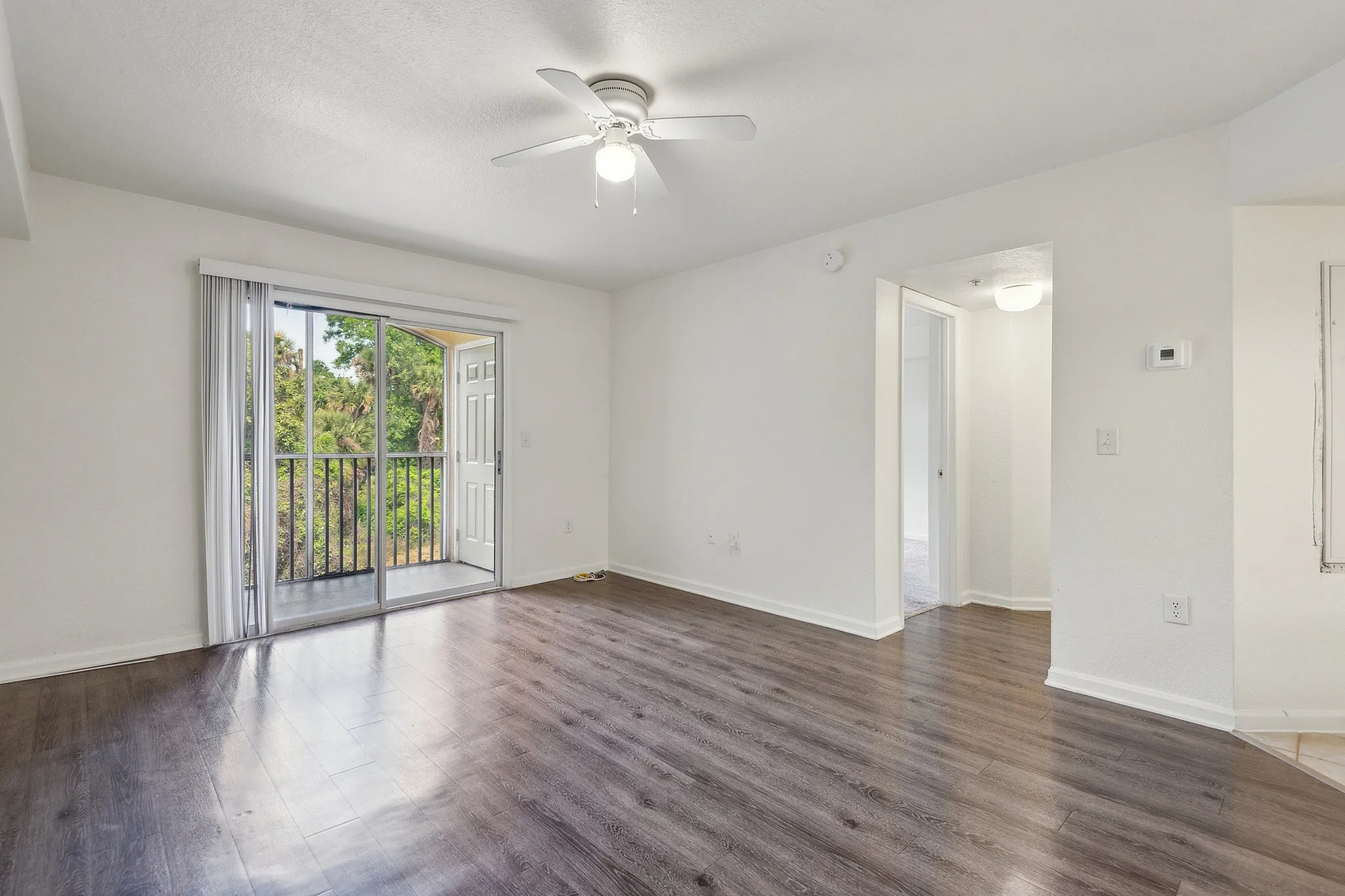 Bright living area with elegant wood flooring, ceiling fan, and sliding glass doors opening to a lush green view.