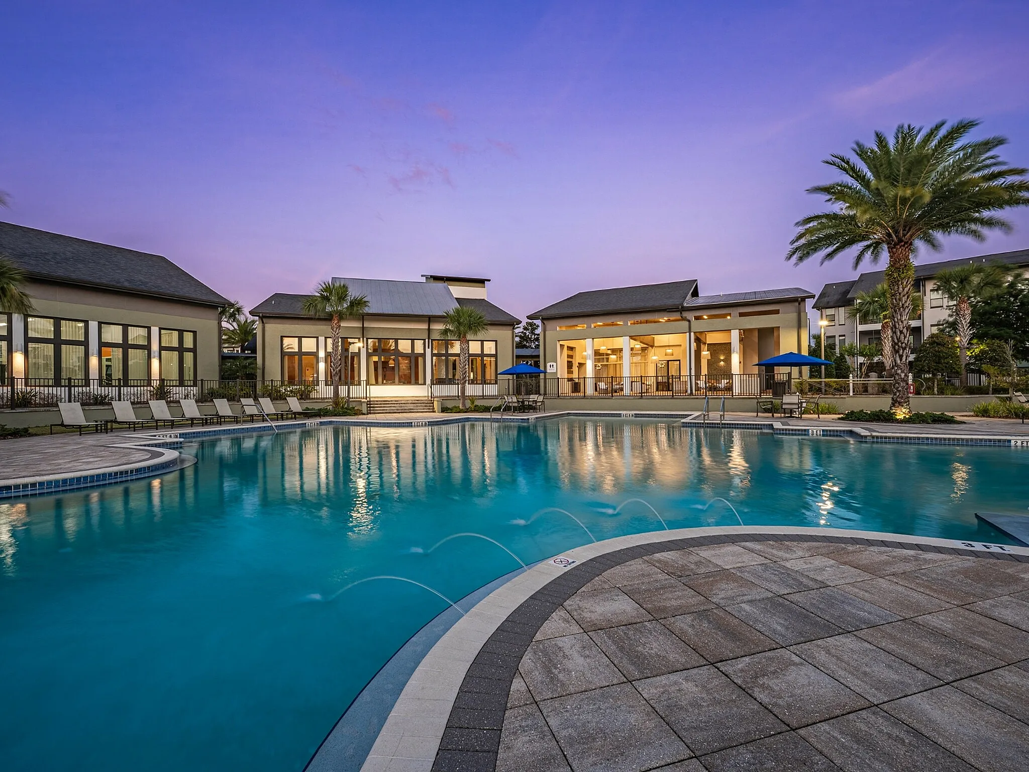 Luxurious apartment complex pool area at sunset with palm trees, modern clubhouse, and inviting lounge chairs.