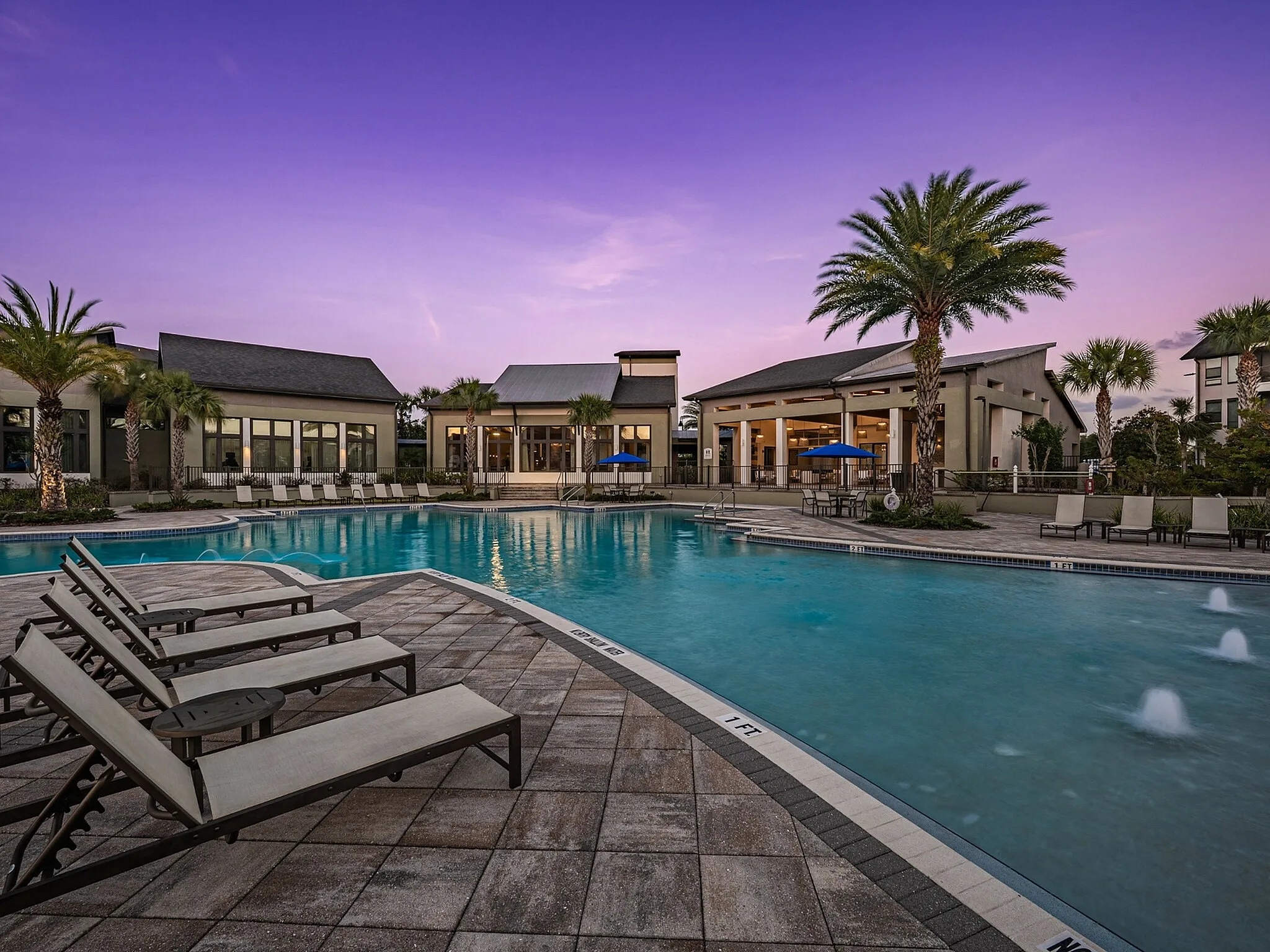 Luxurious apartment complex with a stunning outdoor pool, surrounded by palm trees and sun loungers under a vibrant purple sunset sky.