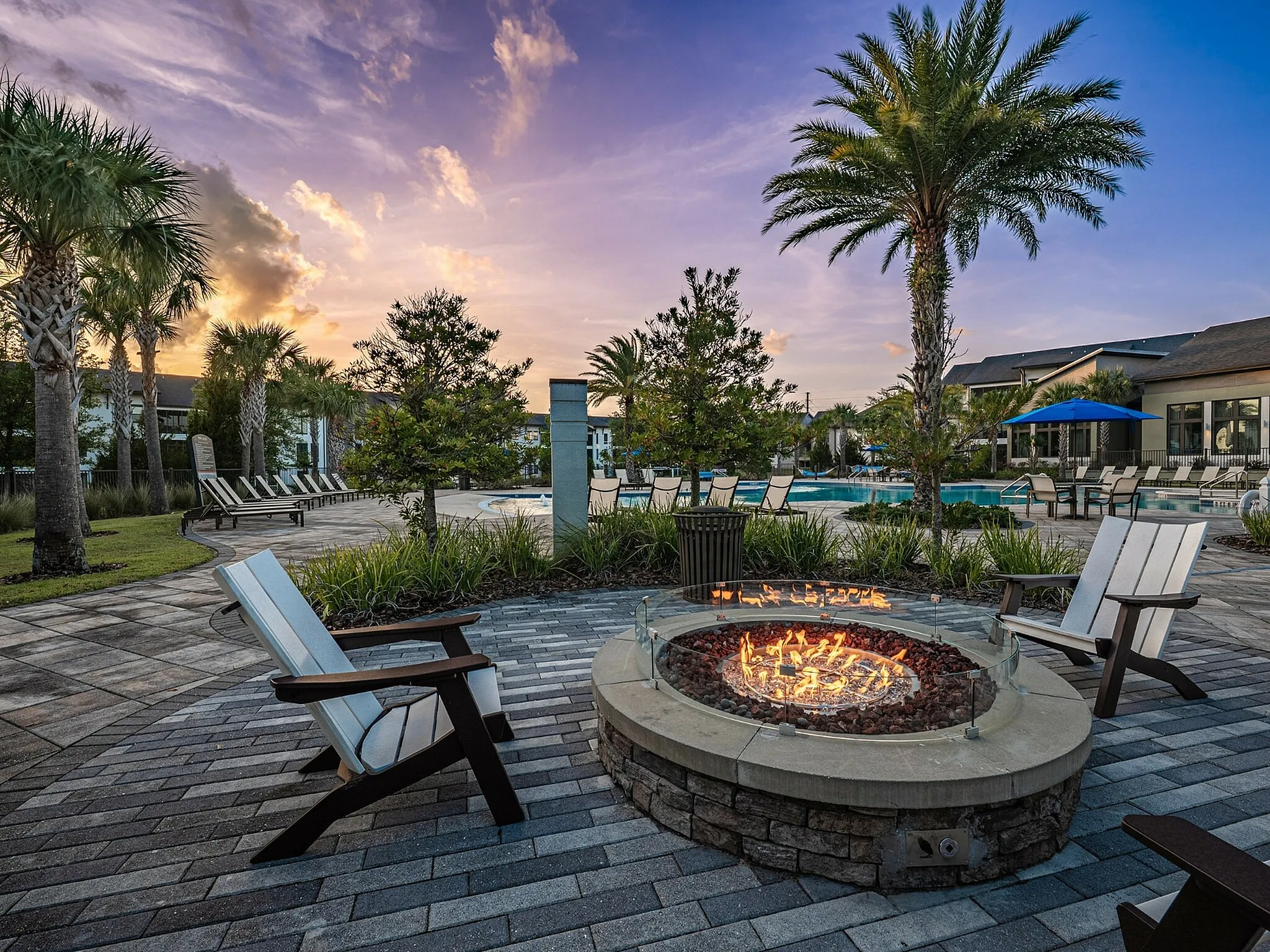 Luxurious outdoor oasis featuring a cozy fire pit surrounded by elegant seating, lush palm trees, and a stunning swimming pool at sunset.
